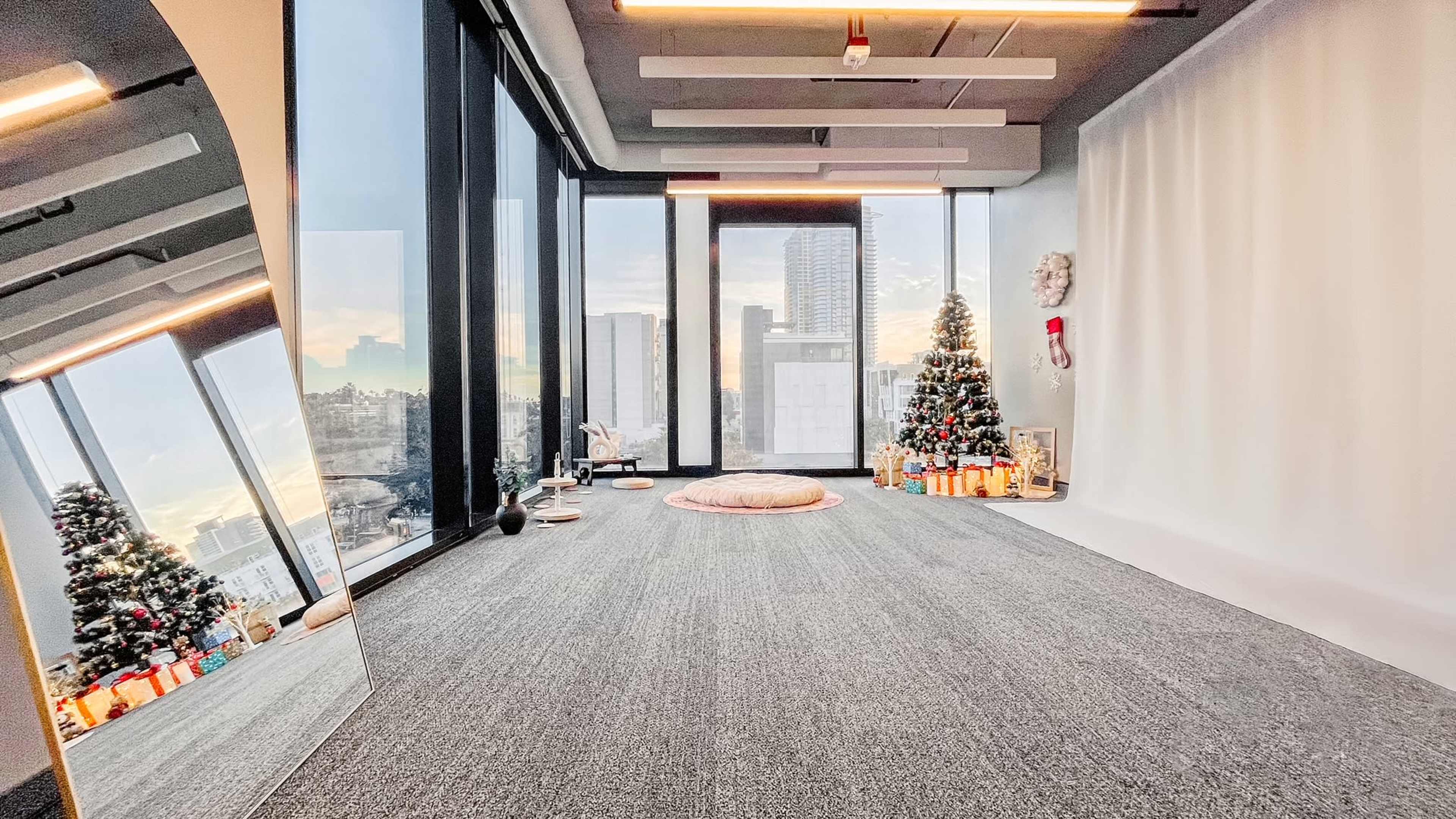 The scene features an empty room with large windows overlooking a cityscape, adorned with a Christmas tree and gifts, and a soft area rug in the center.