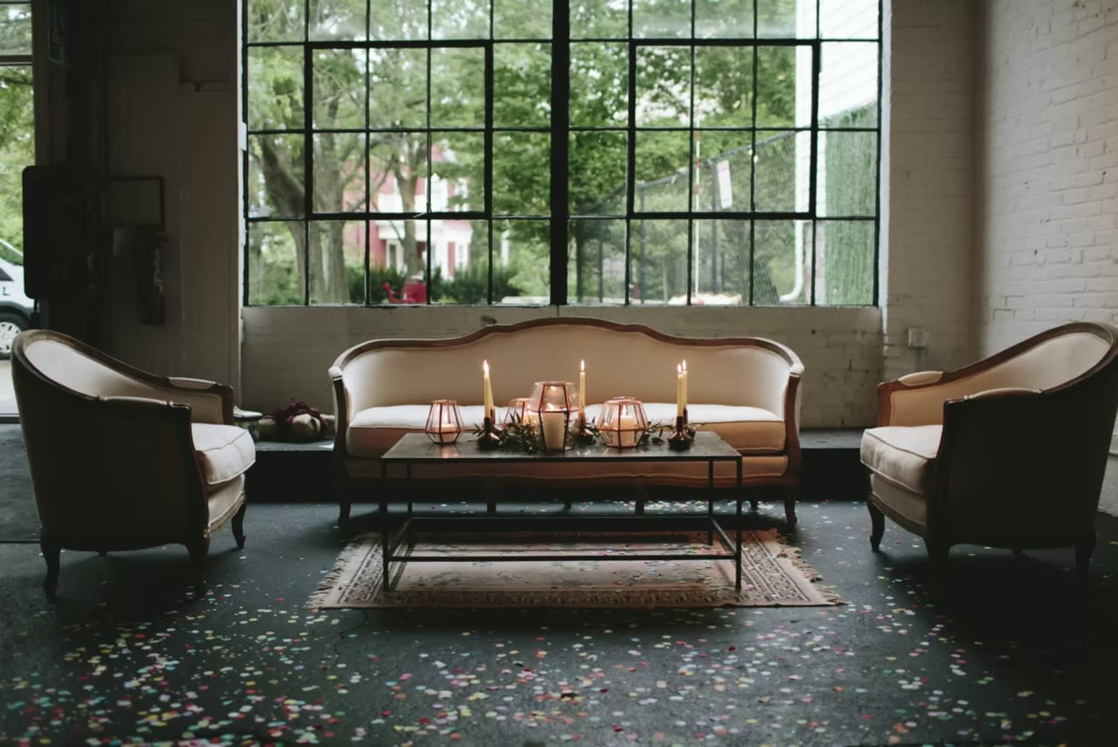 A stylish seating area is set up with a sofa and two chairs around a table, adorned with candles and greenery, in front of large windows letting in natural light.