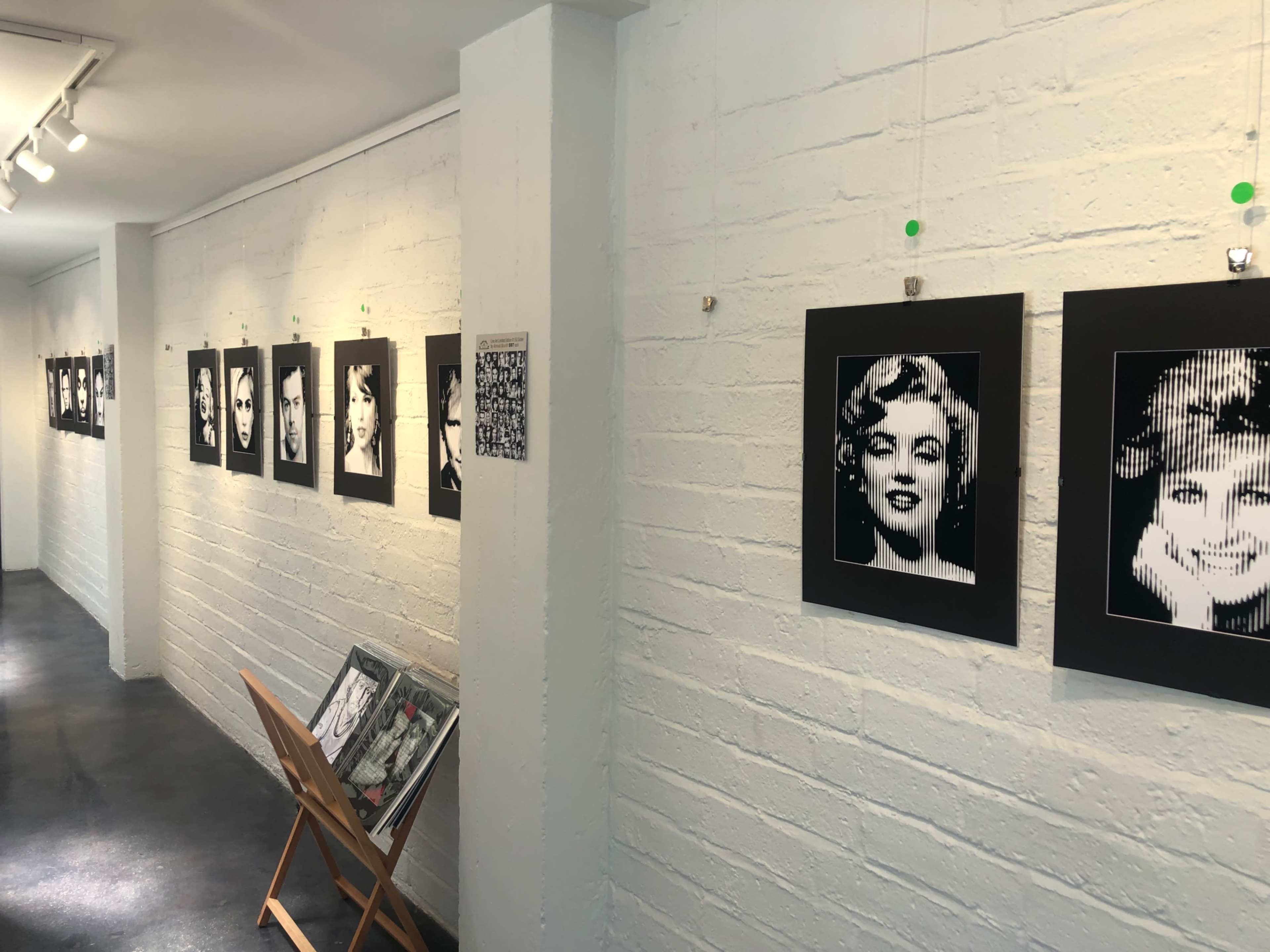 The image shows an art gallery hallway with a series of black and white portraits hanging on white brick walls, along with a display of art materials on a wooden easel.