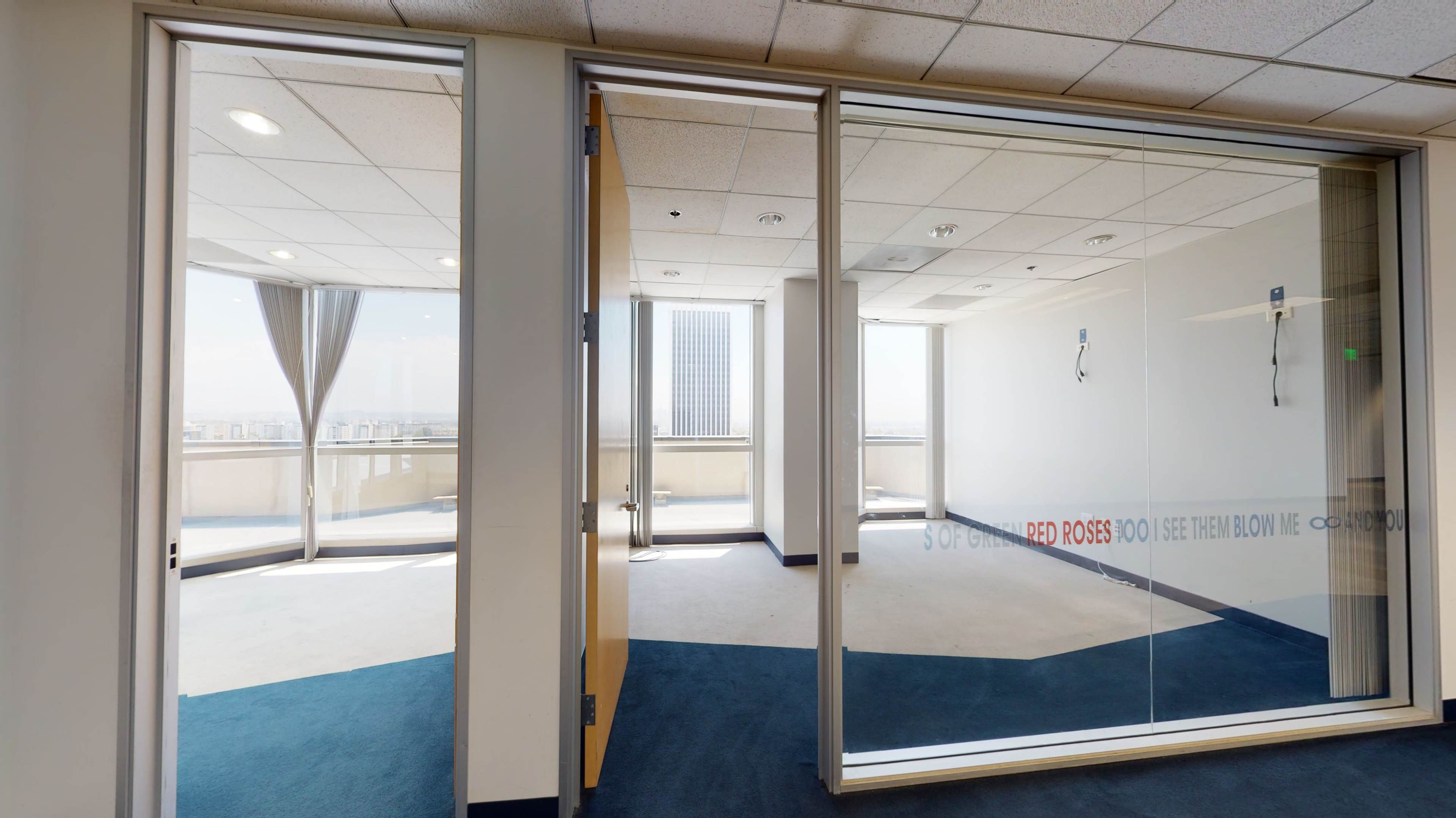 The image shows a modern office space with glass walls, featuring an empty room with blue carpet and large windows overlooking a cityscape.