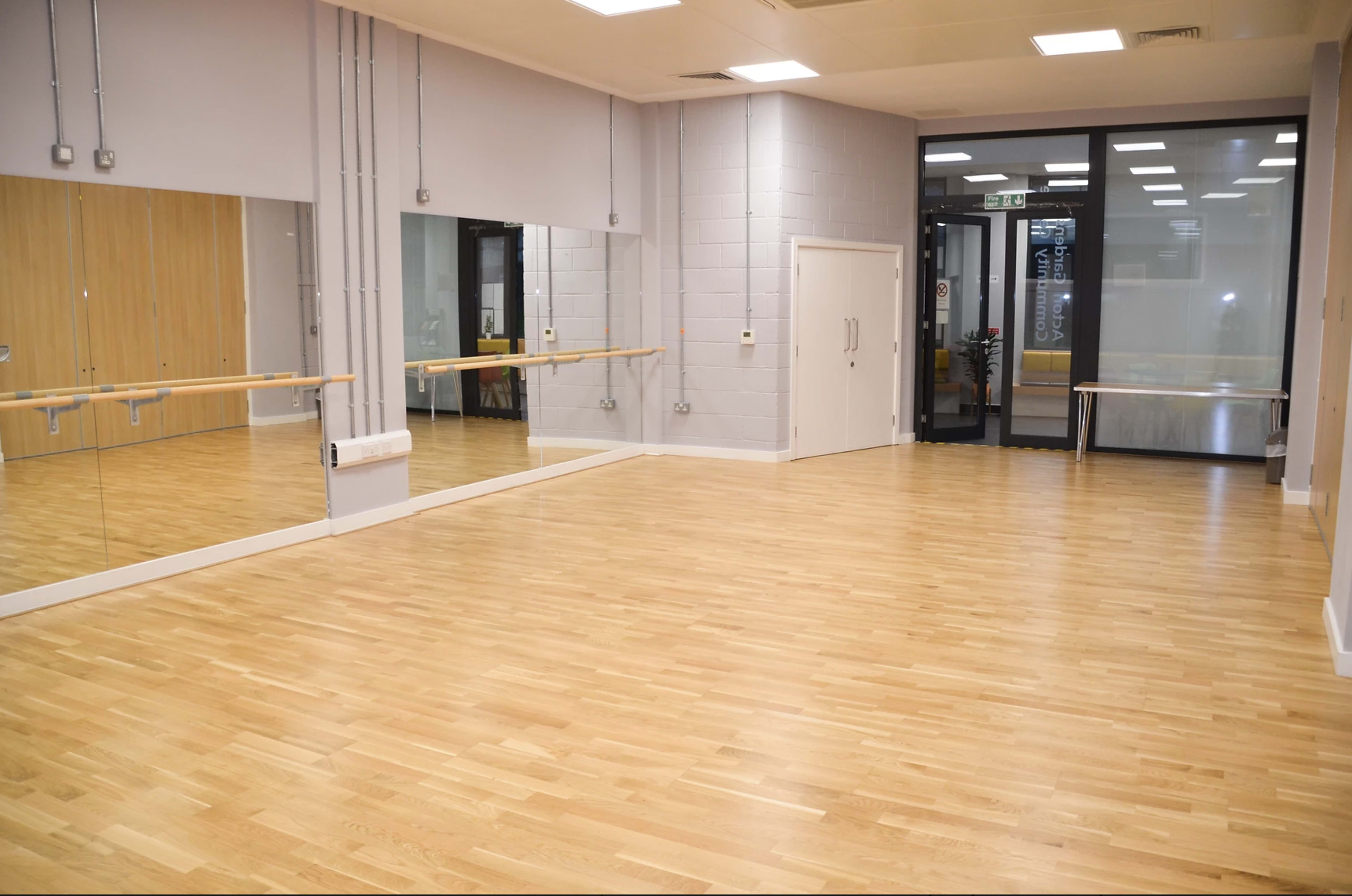 A spacious wooden-floored studio with mirrored walls and an entrance area featuring a glass door.