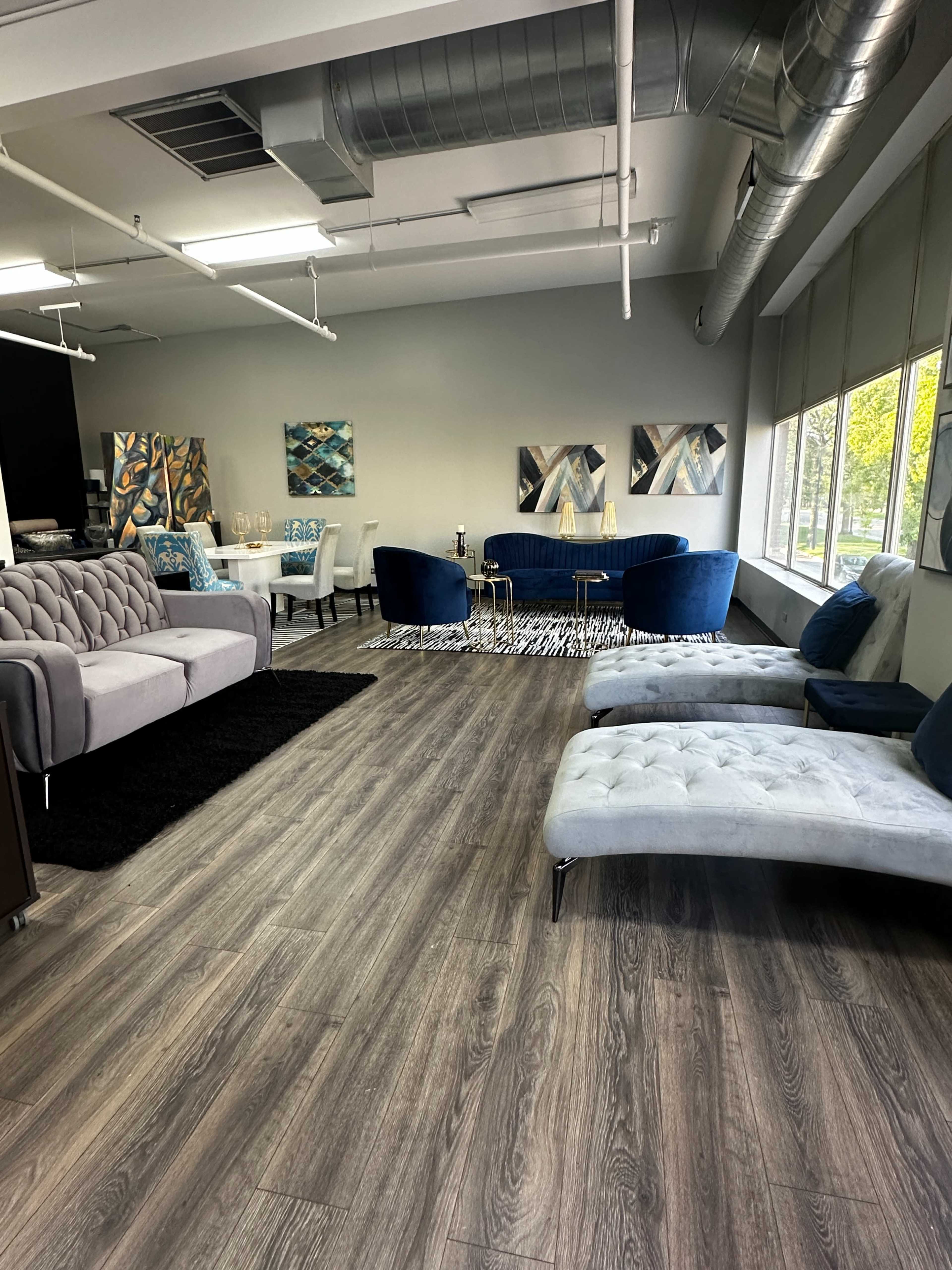 The image shows a modern lounge area with gray and blue furniture, wooden flooring, and large windows allowing natural light.