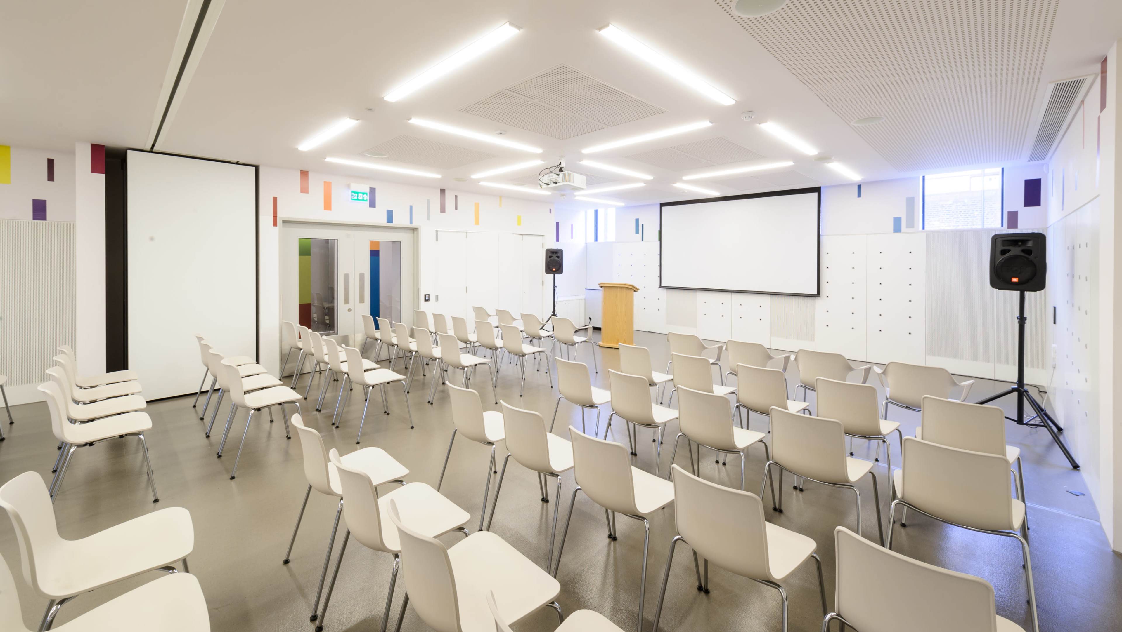 The image shows a modern conference room set up with rows of white chairs facing a wooden podium and a large screen.
