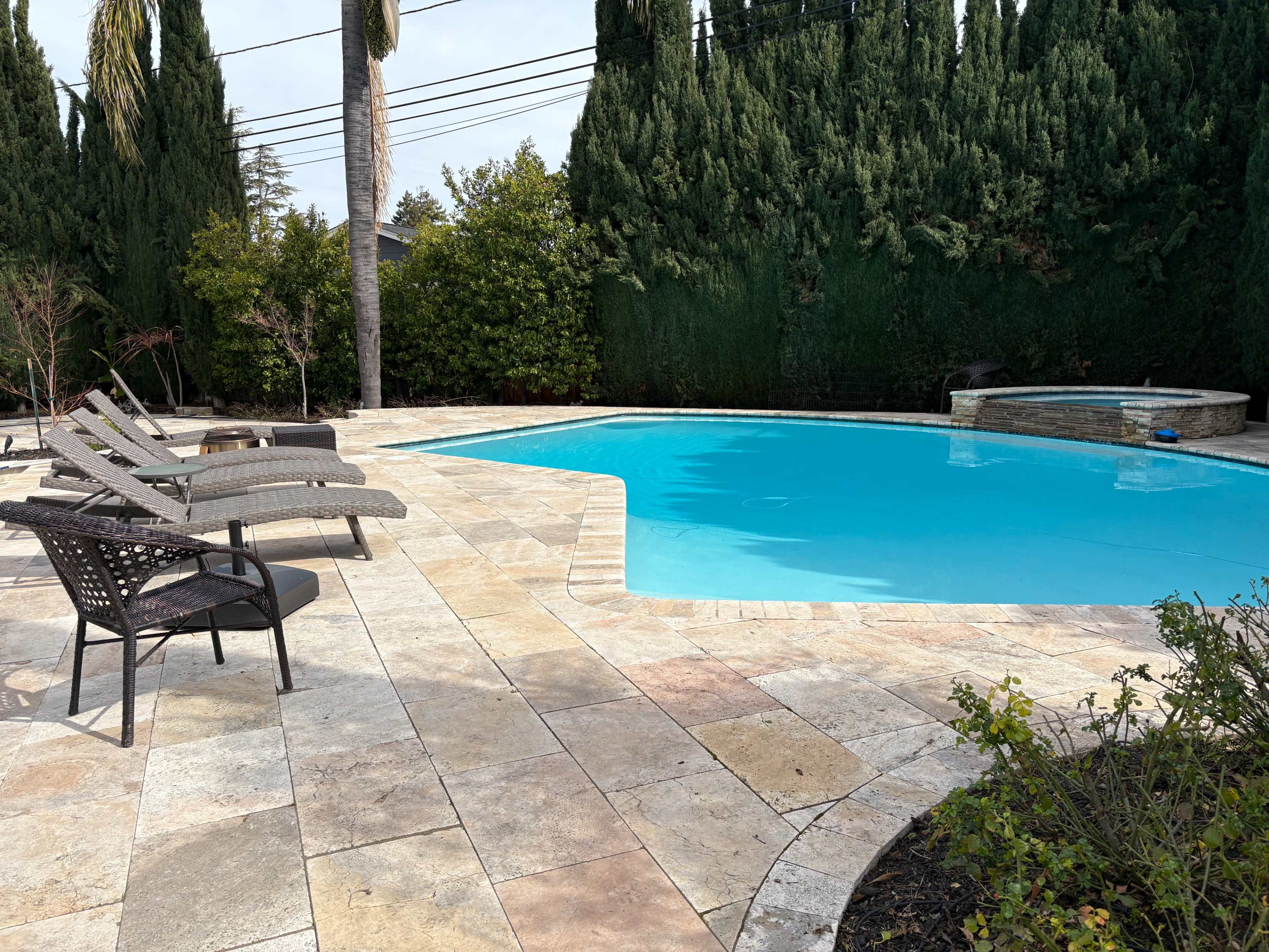 A clear blue swimming pool is surrounded by stone paving and lounge chairs, with tall evergreen trees in the background.