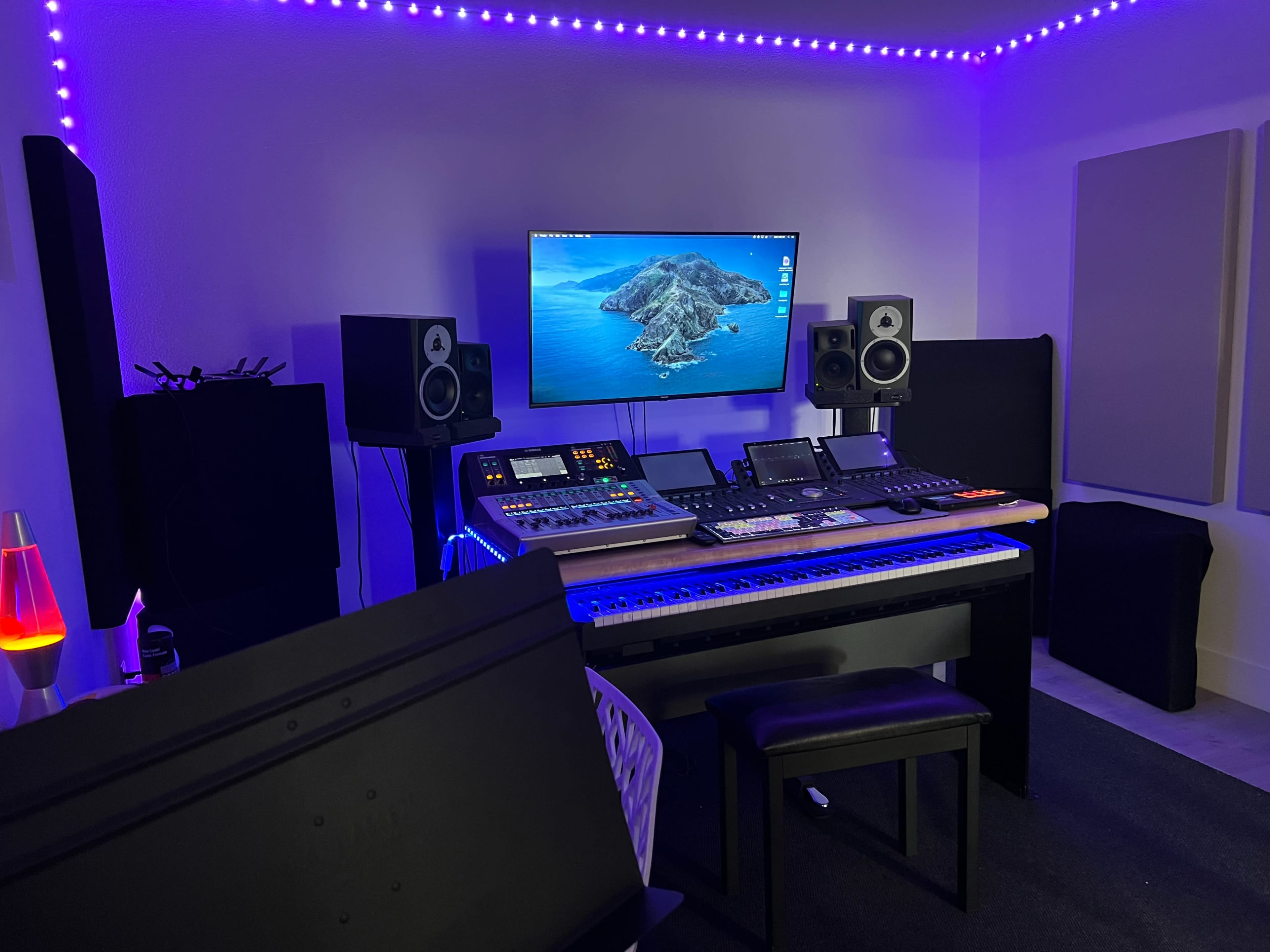 A modern music studio is equipped with a computer monitor, mixing console, speakers, and a keyboard, illuminated by purple LED lights.