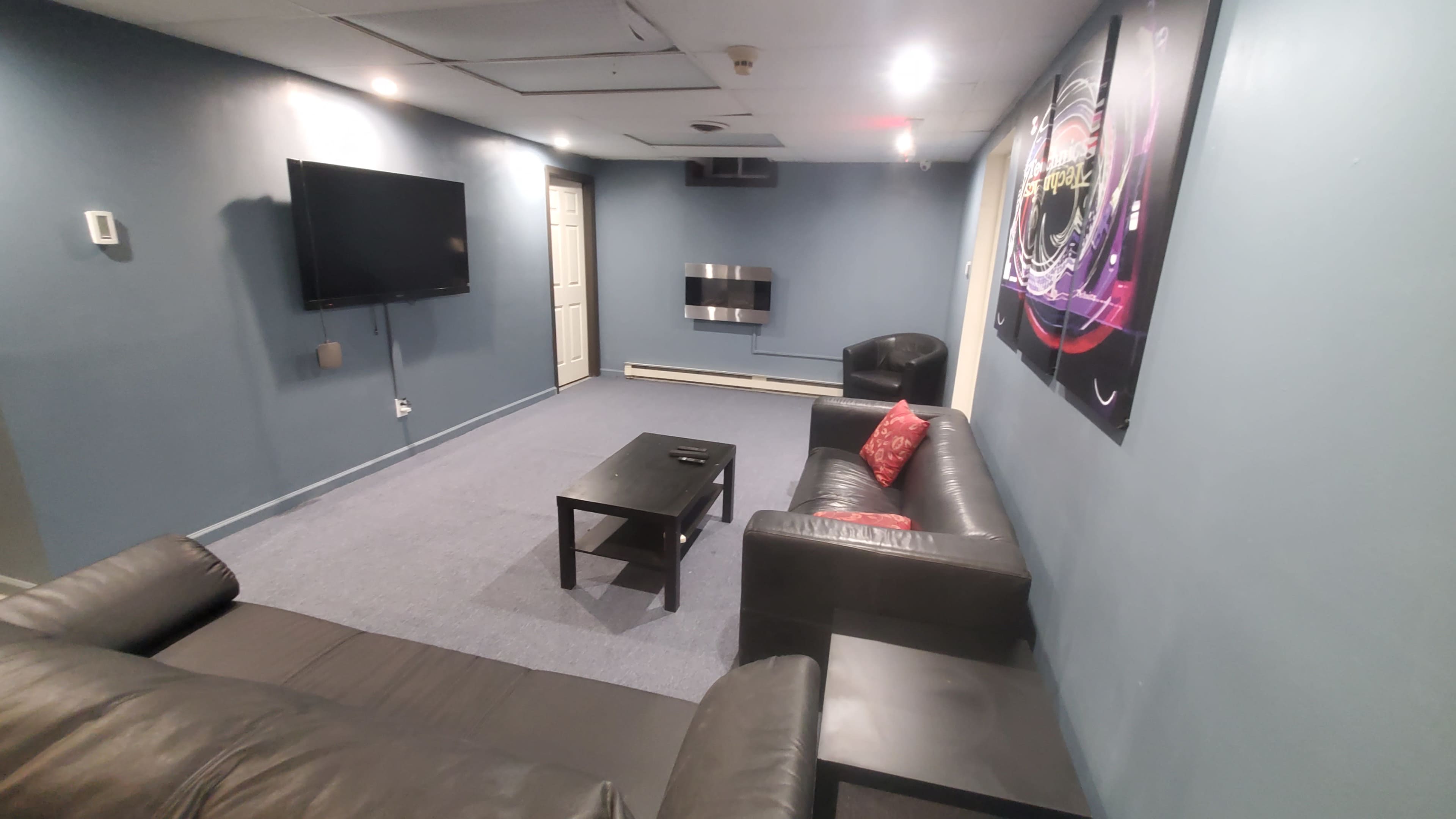 A sparsely furnished room with a large flat-screen TV, a wall-mounted fireplace, two black leather chairs, a black coffee table, and blue walls.