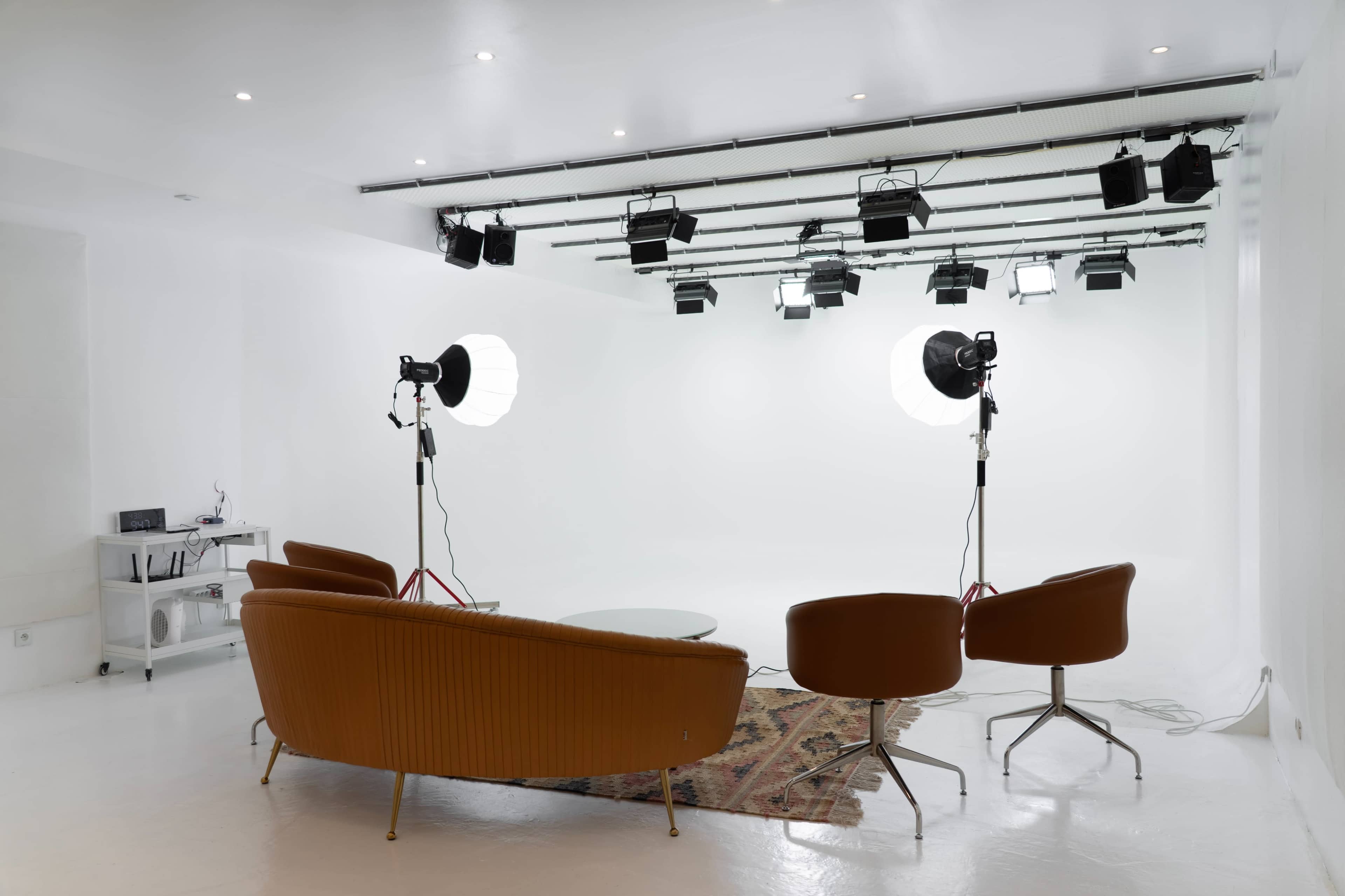 A minimalist photography studio features two lighting setups and a mid-century modern seating area on a white backdrop.
