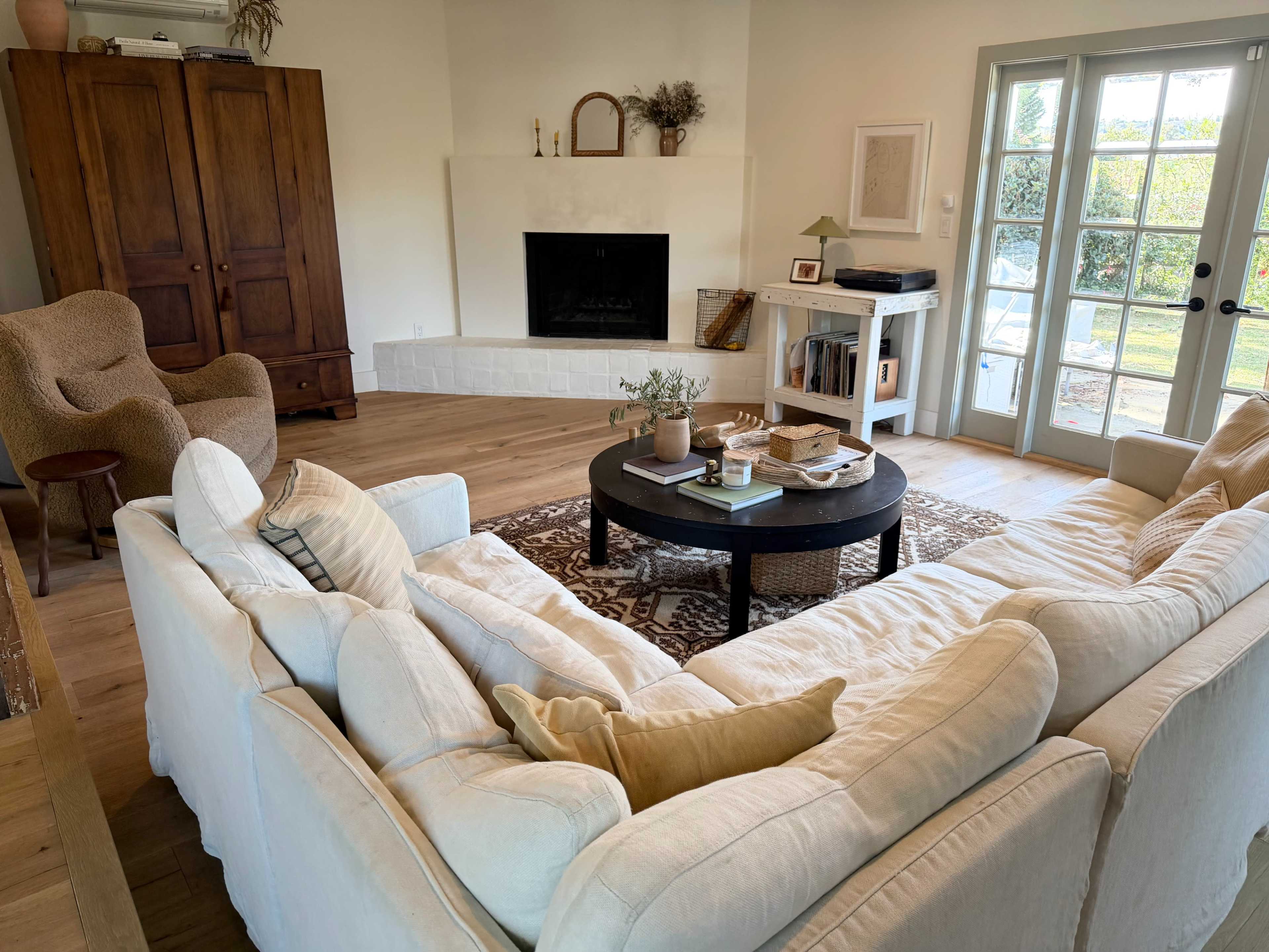 The image shows a cozy living room featuring a large white sectional sofa, a round coffee table with books and plants, a fireplace, and large glass doors leading to an outdoor area.