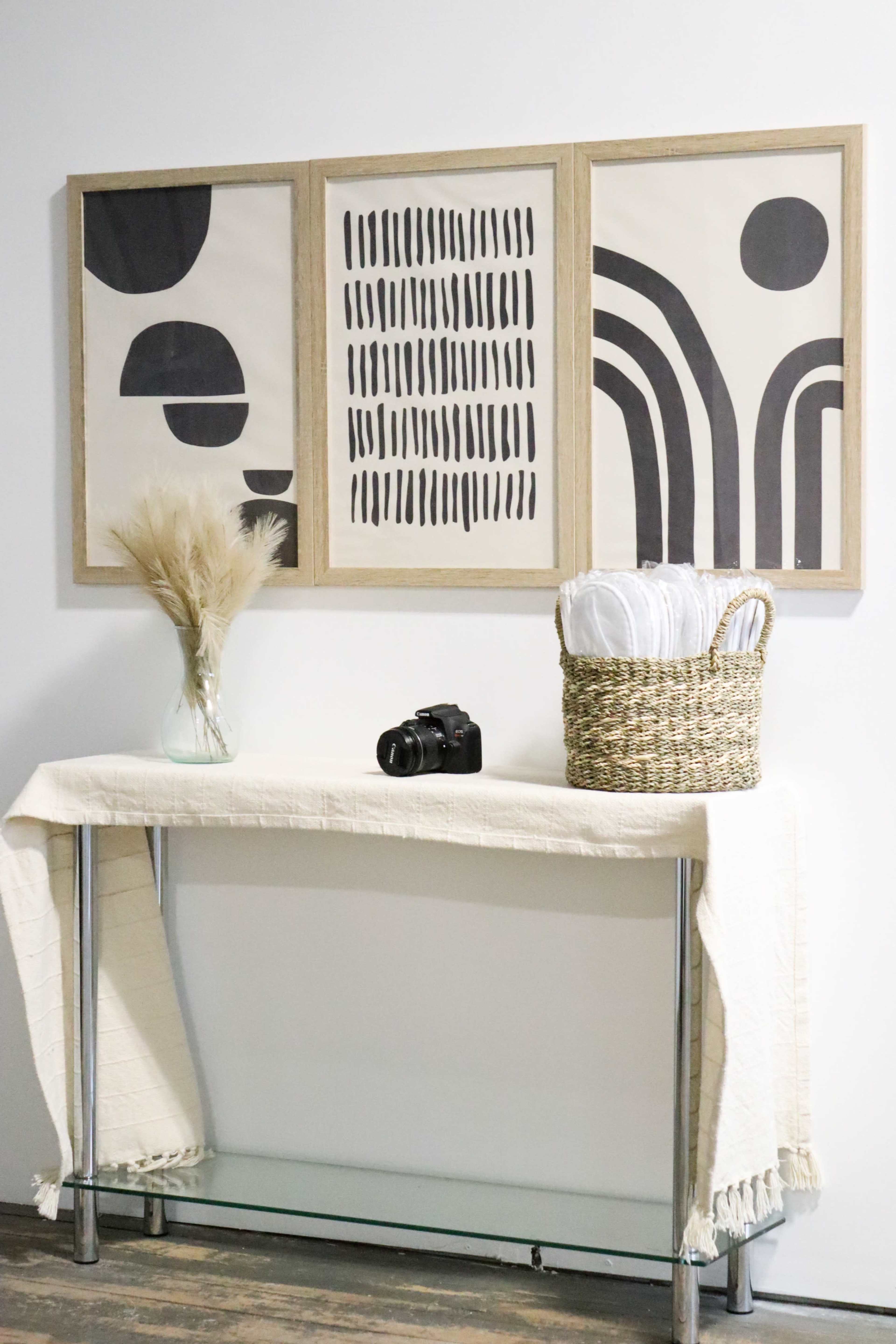 A console table is displayed against a wall featuring three framed art pieces and a vase with dry foliage, accompanied by a camera and a woven basket.