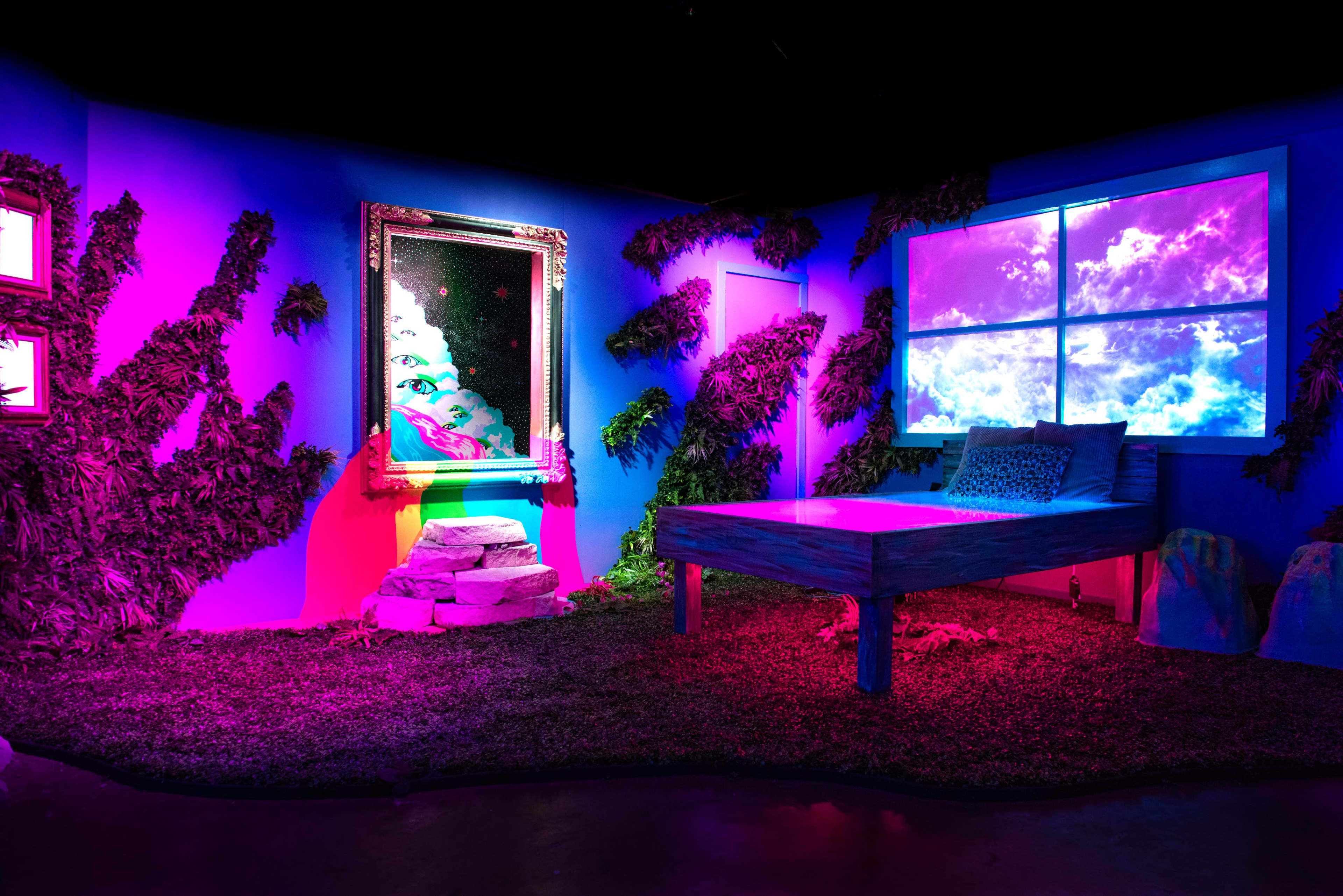 The image depicts a vibrant, art installation featuring a bed, a large mirror, and a window with a colorful sky scene, all surrounded by green foliage and dramatic lighting.