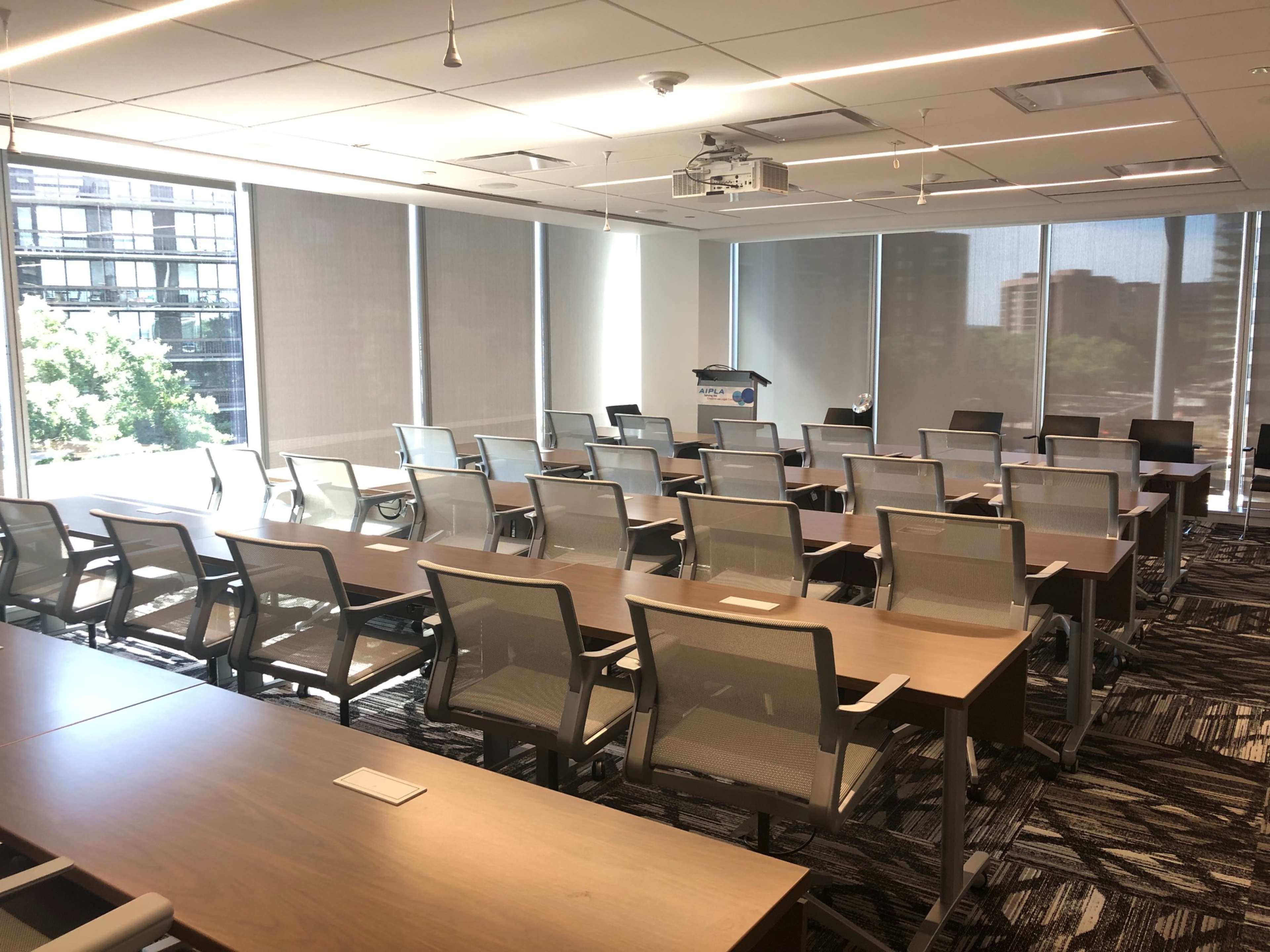 An empty conference room with rows of chairs arranged around wooden tables, large windows letting in natural light, and a projector mounted on the ceiling.