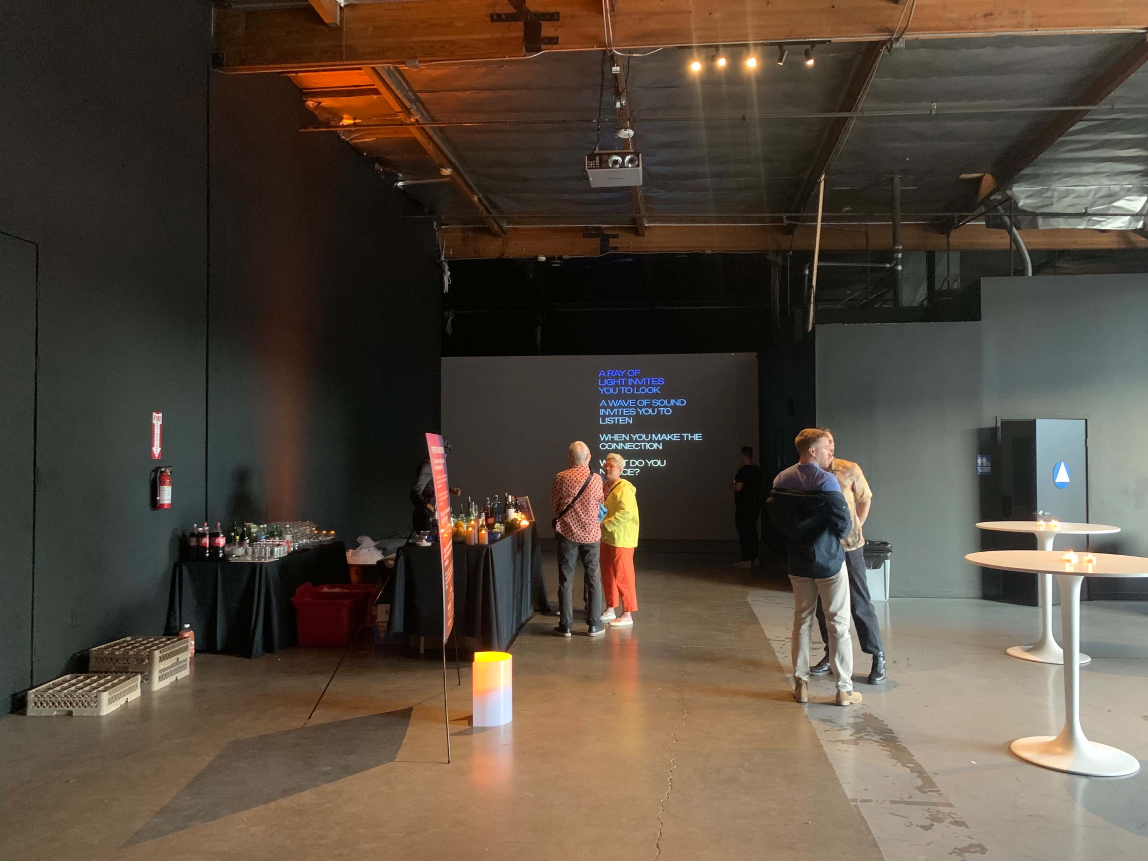 A group of people gather at a dimly lit event space with drinks on a table and a large screen displaying a message.