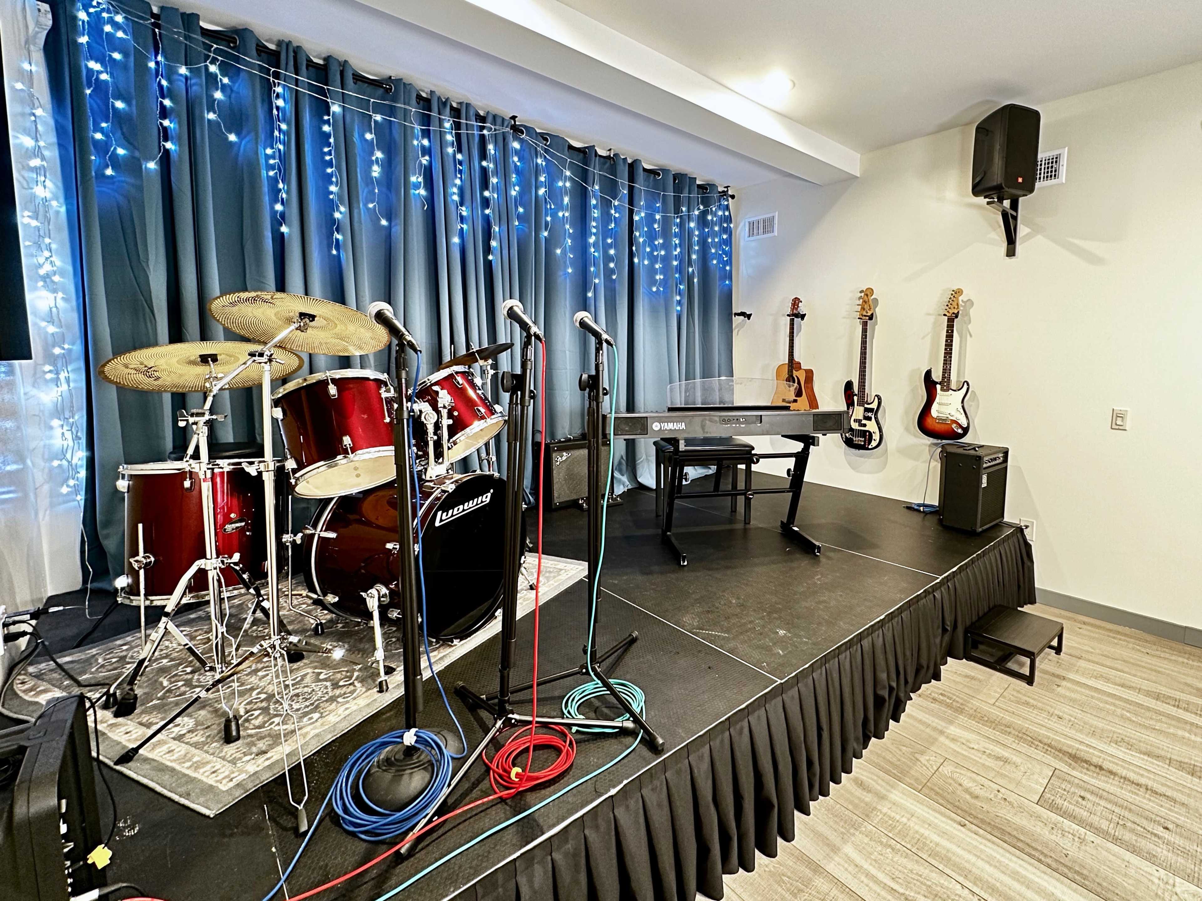 A small stage setup features a drum kit, keyboard, and various electric guitars on the wall, surrounded by blue curtain and string lights.