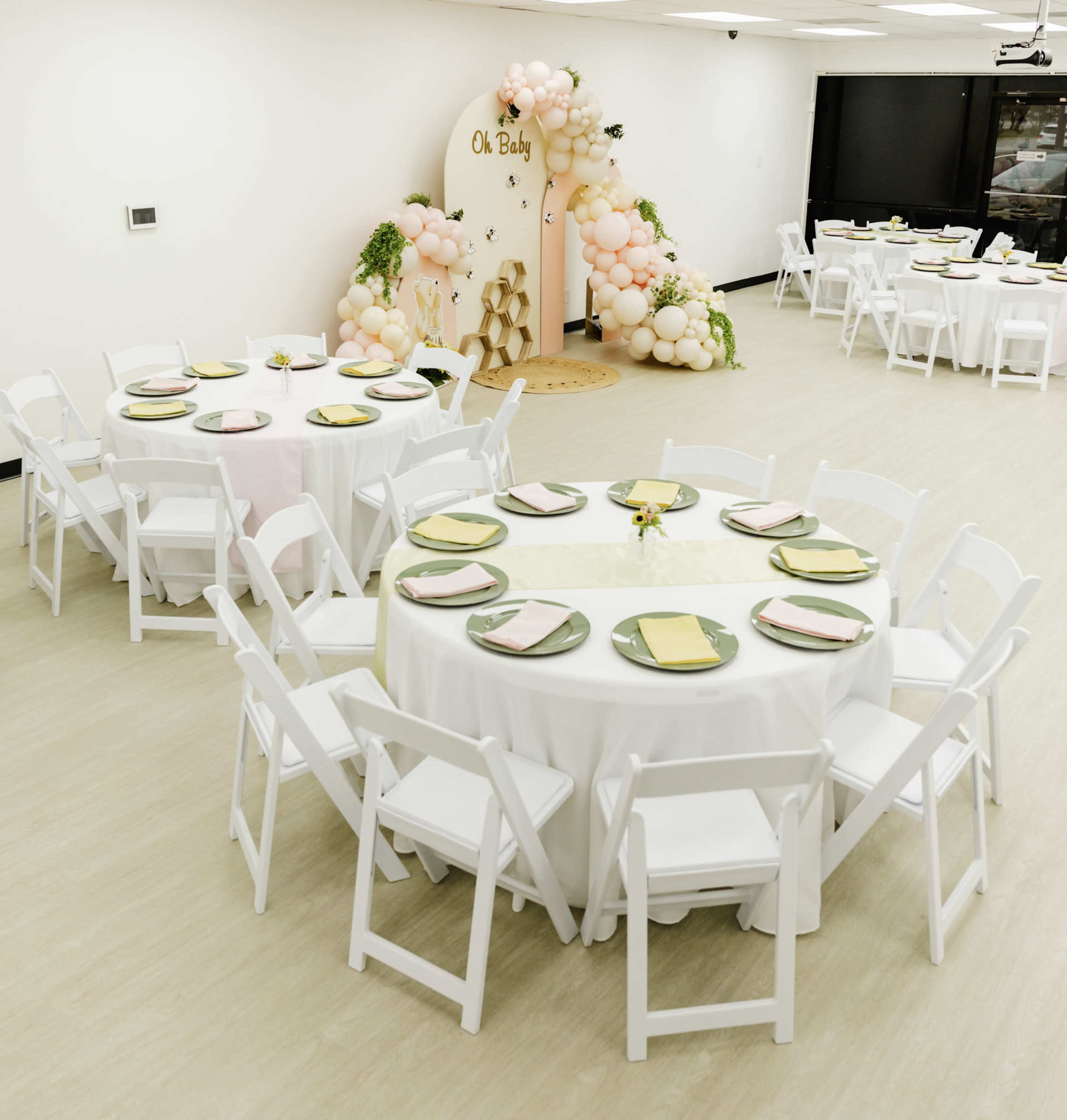 The image shows a spacious event room with two circular tables set for a gathering, featuring white chairs, pastel-colored plates, and a decorative backdrop of balloons and a sign that reads "Oh Baby."