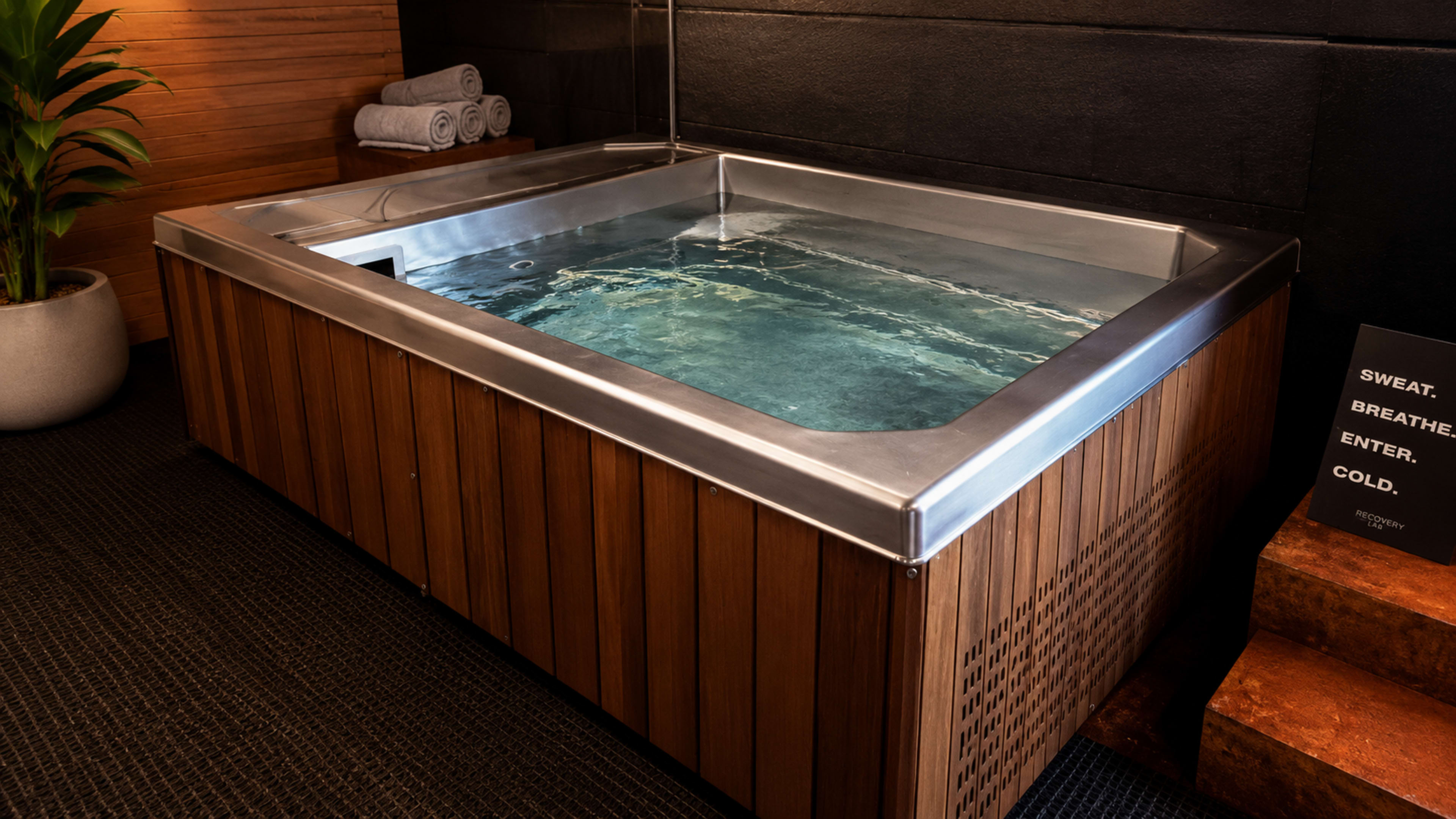 The image shows a wooden hot tub filled with water, positioned in a modern spa-like setting with neatly stacked towels nearby.
