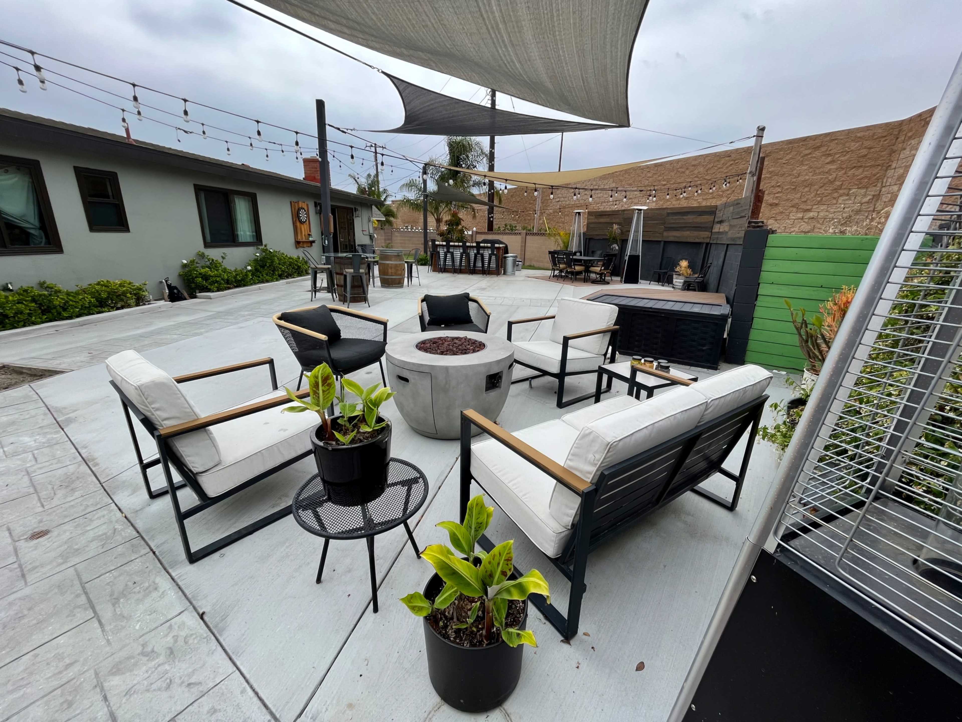 A modern outdoor seating area with several chairs, a central fire pit, and shade sails overhead, surrounded by green plants and a barbecue area in the background.