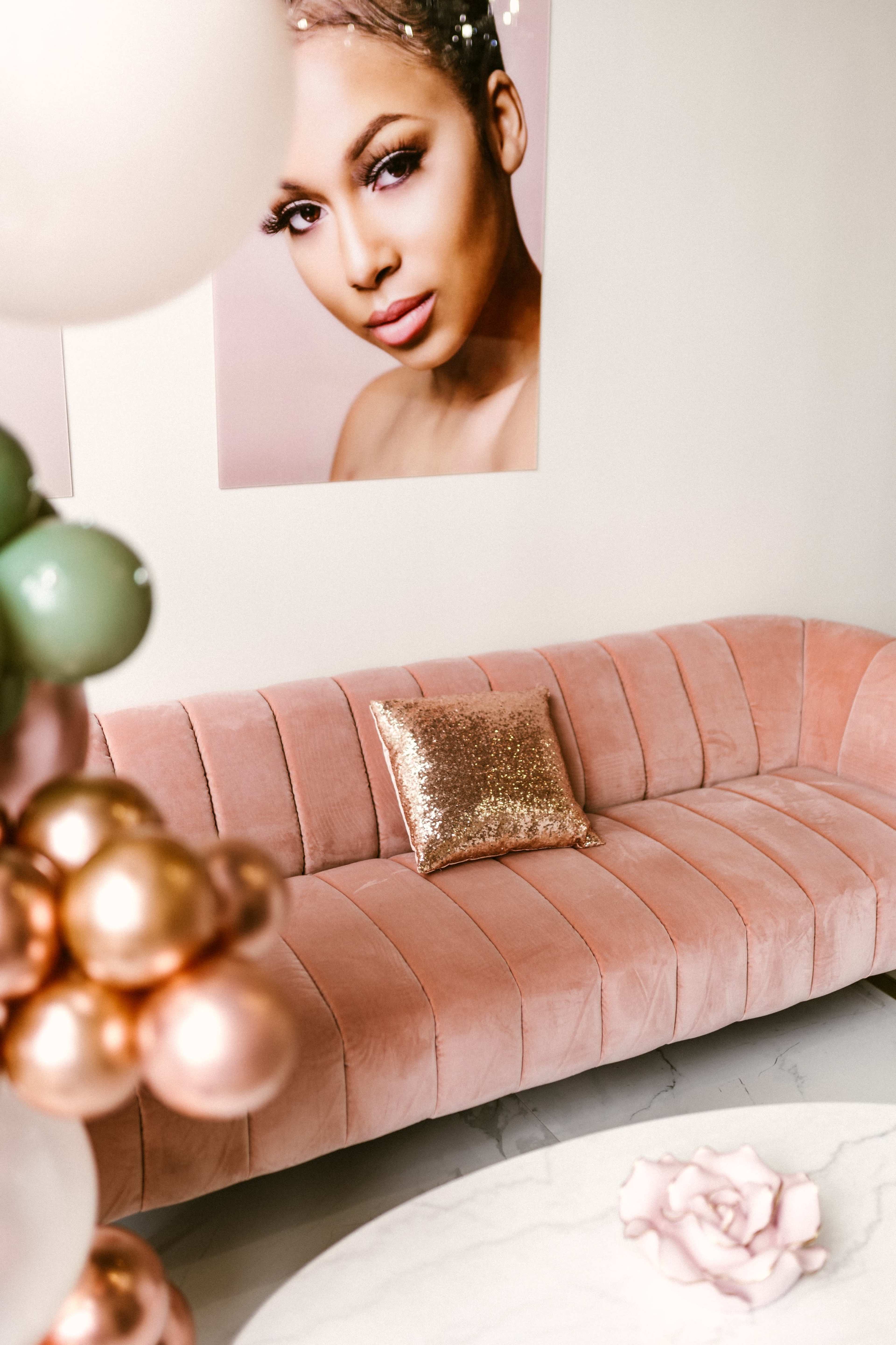 A plush, pink velvet sofa with a sparkling cushion sits against a wall adorned with a large portrait, beside a decorative arrangement of colorful balloons.