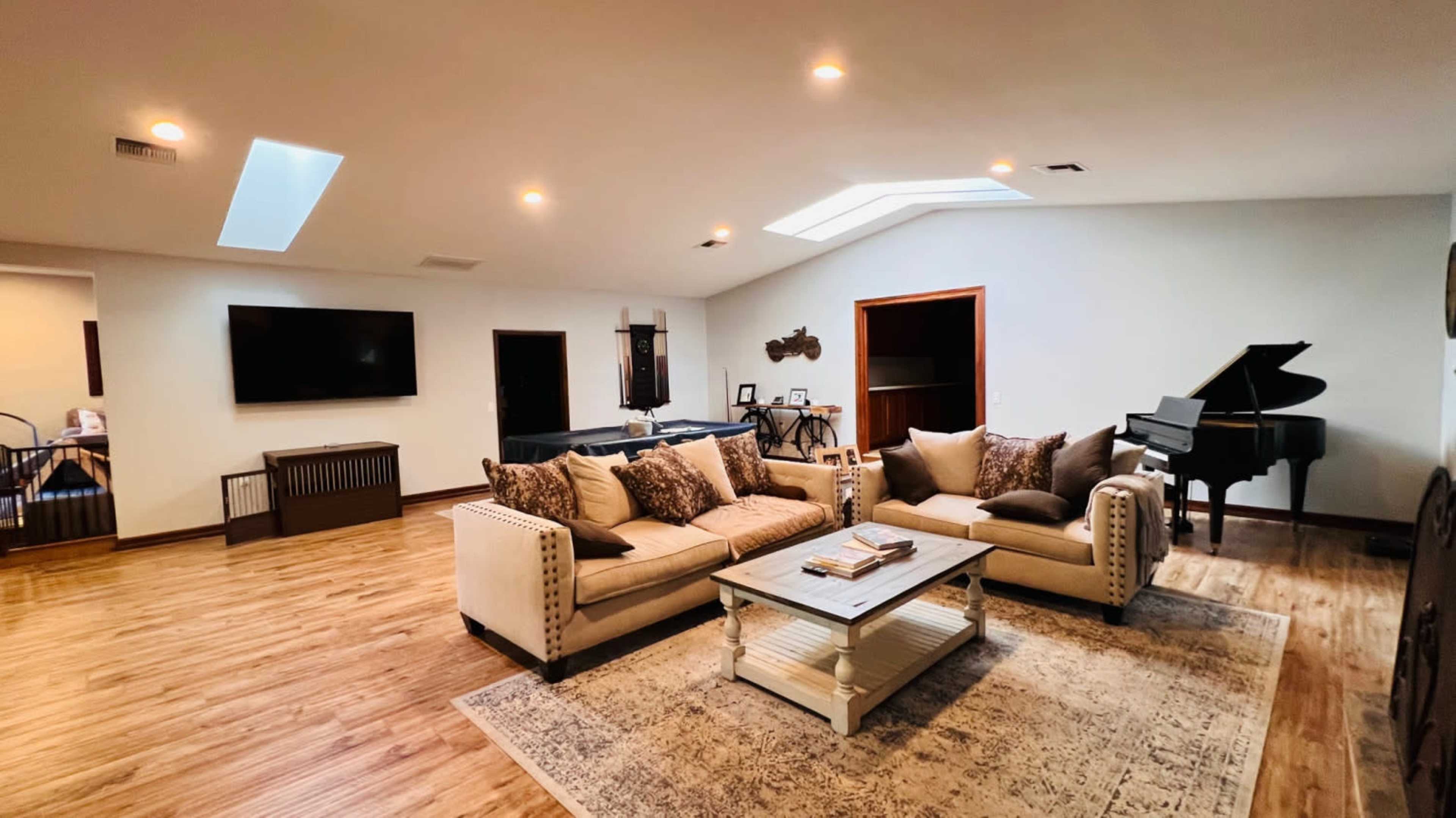A spacious living room features a tan sectional sofa, a light coffee table, and a grand piano, with a skylight illuminating the wooden floor.