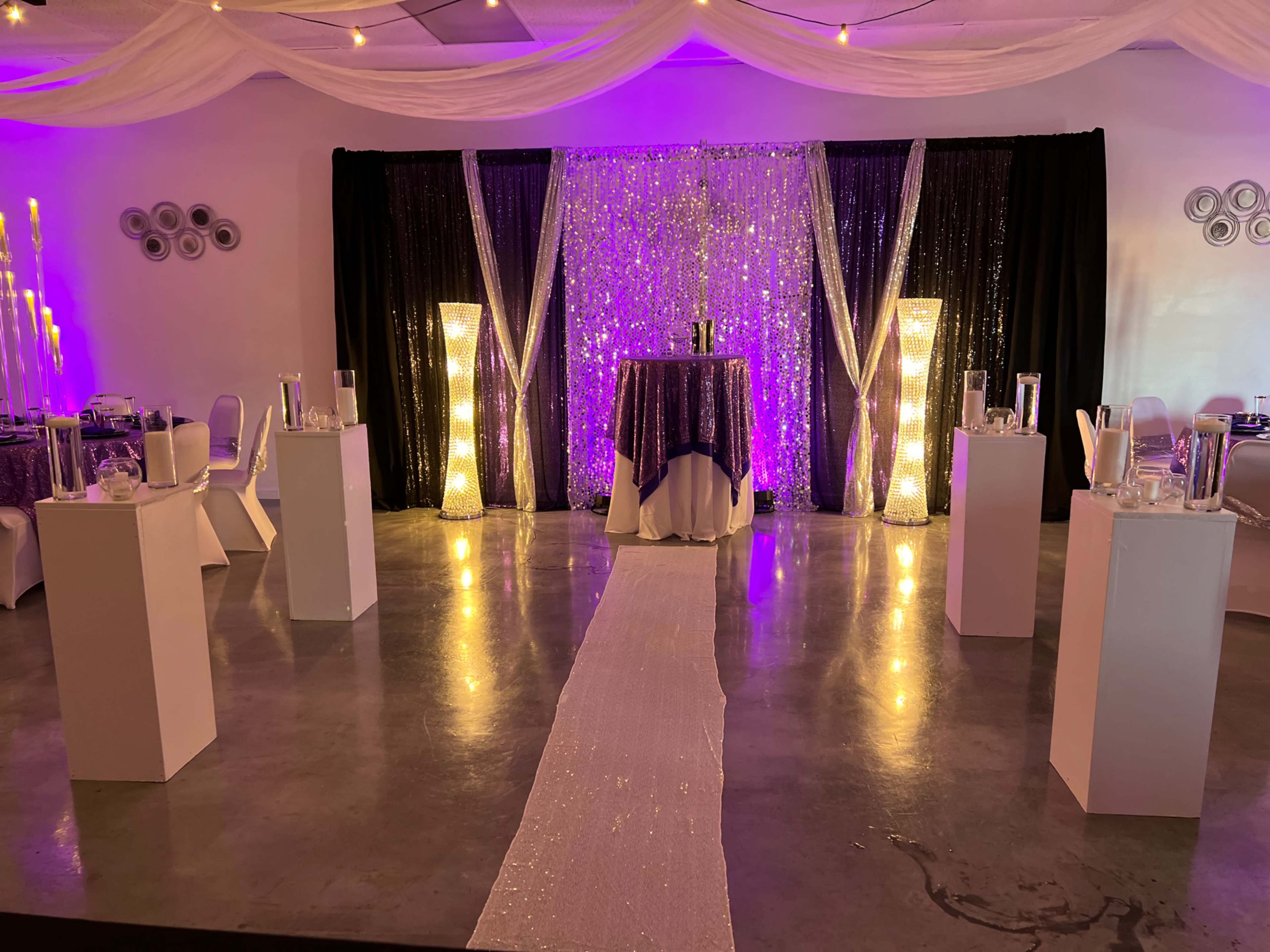 A banquet hall decorated with sparkling backdrops, tall illuminated pillars, and a white runner leading to a central table.