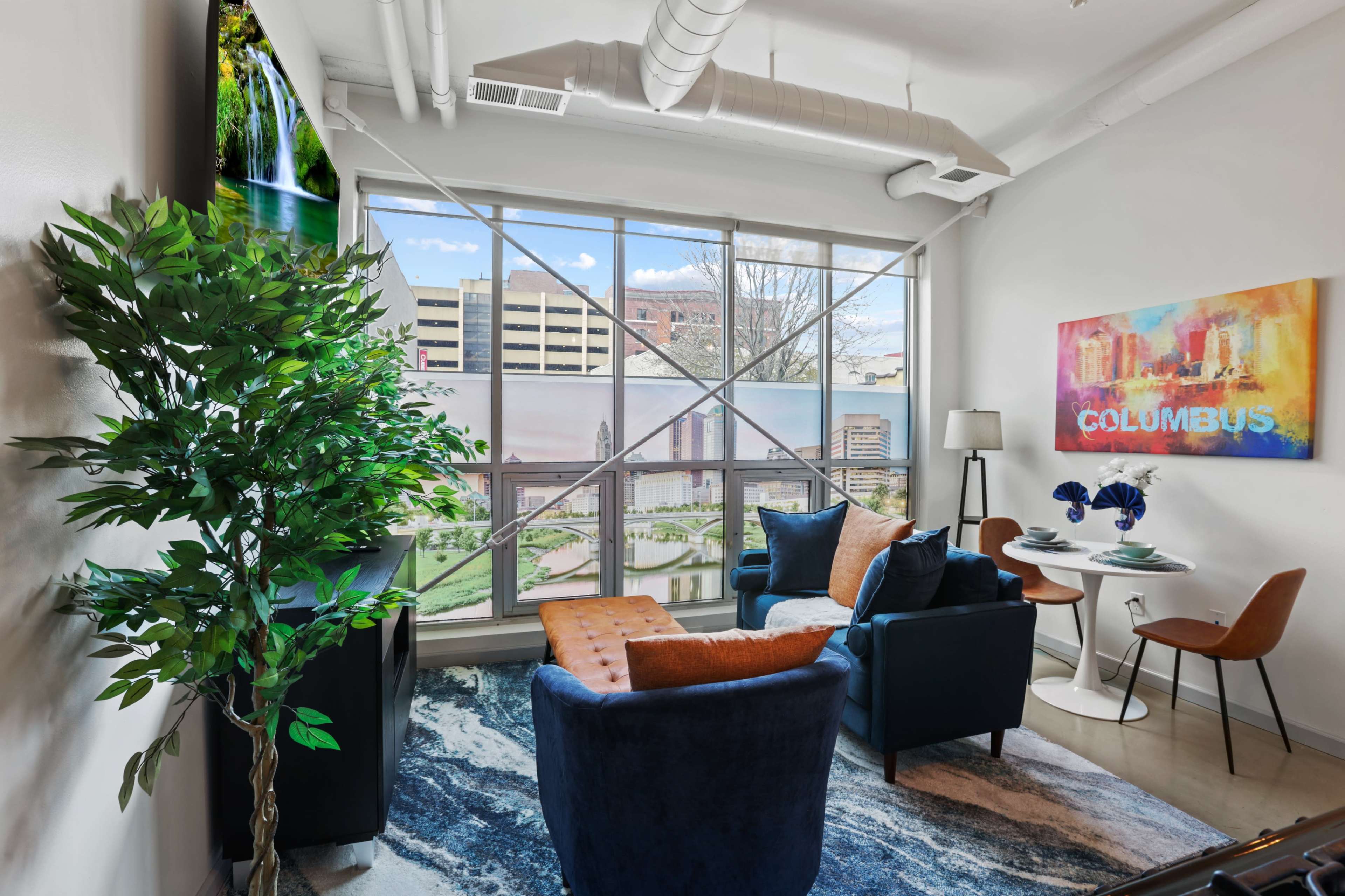 A cozy living area features a blue armchair, an orange ottoman, and a small dining table, with large windows showcasing a cityscape view.