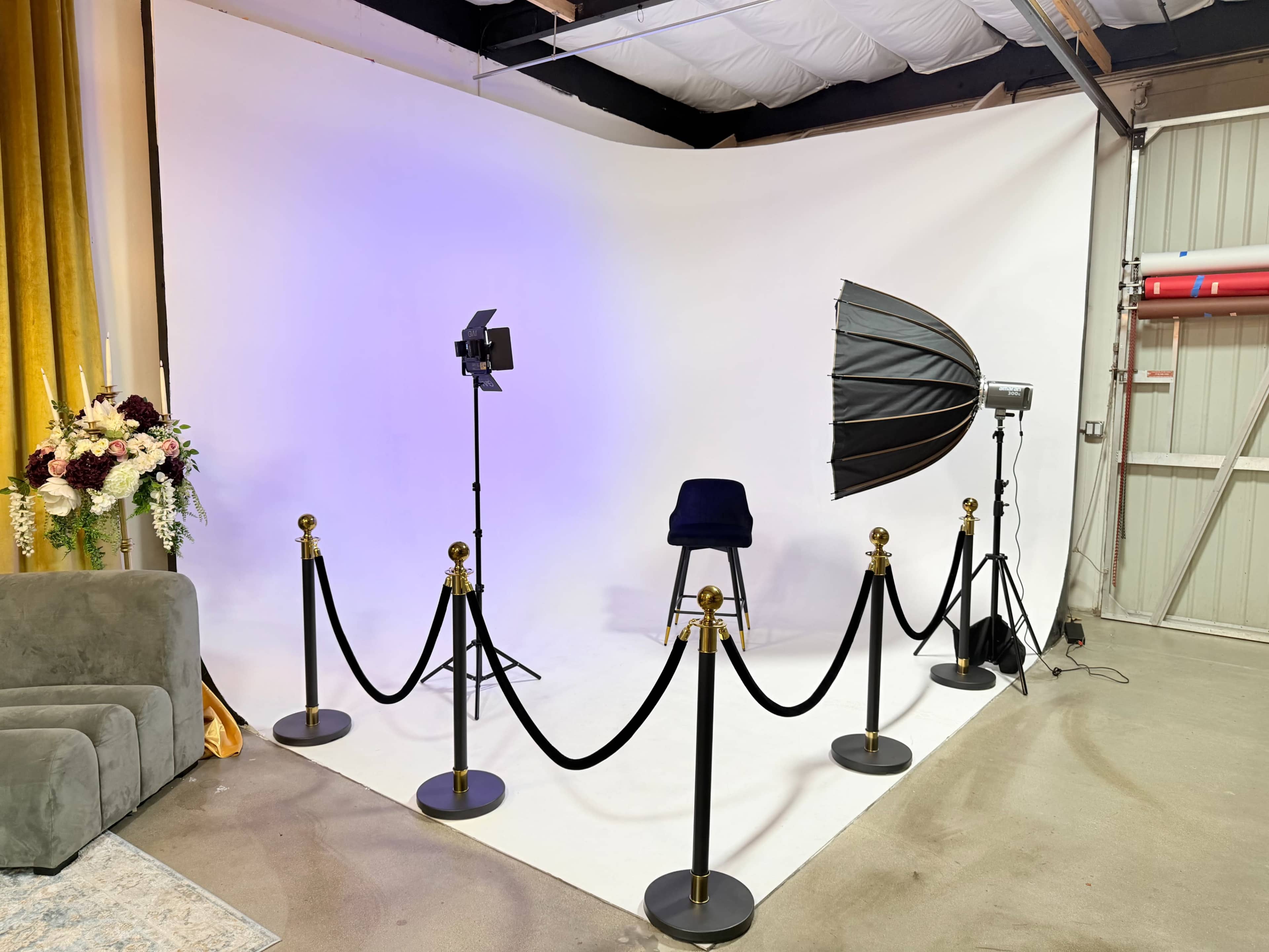 The image shows a photography studio setup with a white backdrop, a chair, lighting equipment, and a velvet rope barrier.