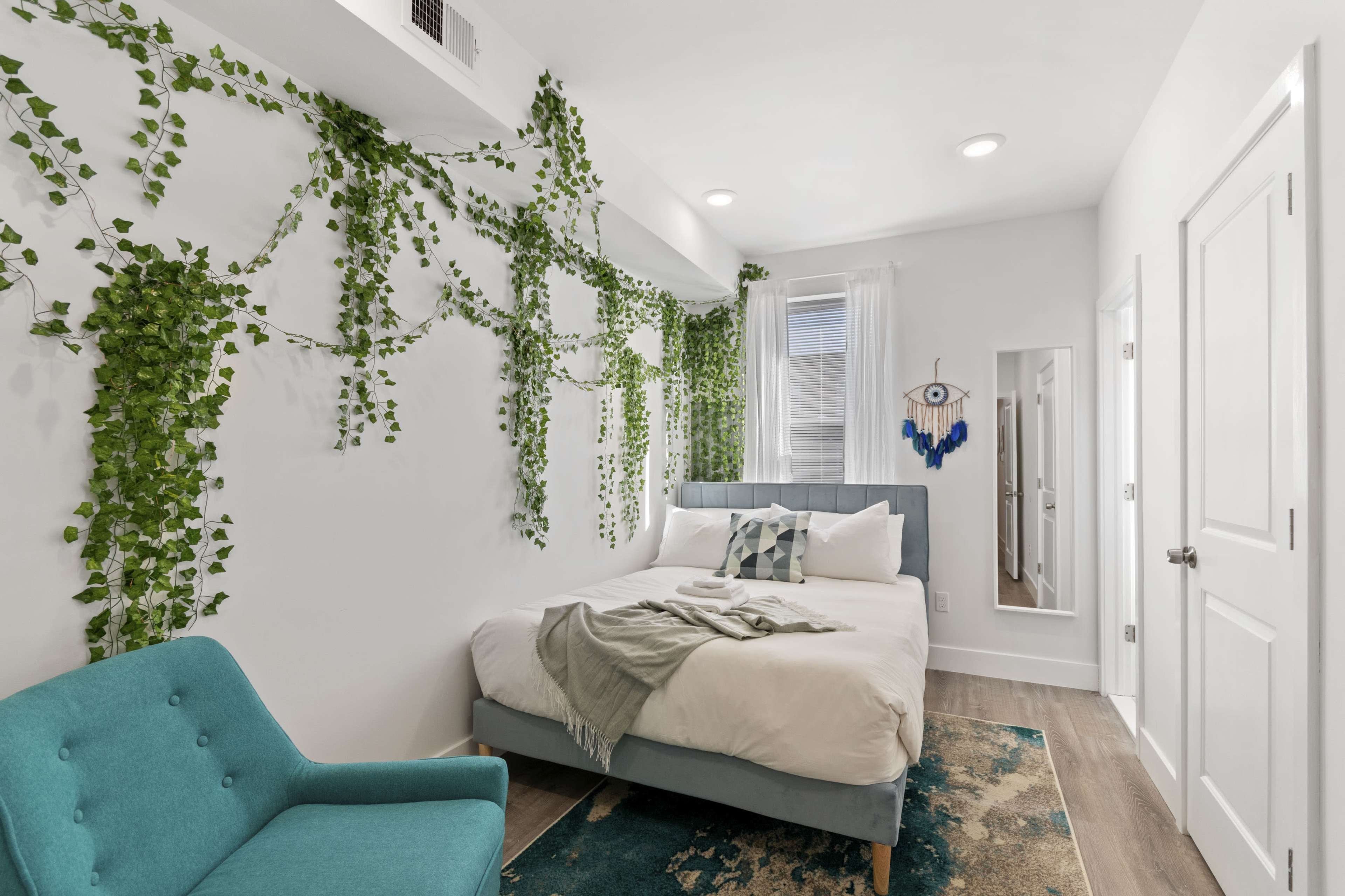 A compact bedroom with a bed, turquoise chair, and decorative greenery on the white walls.