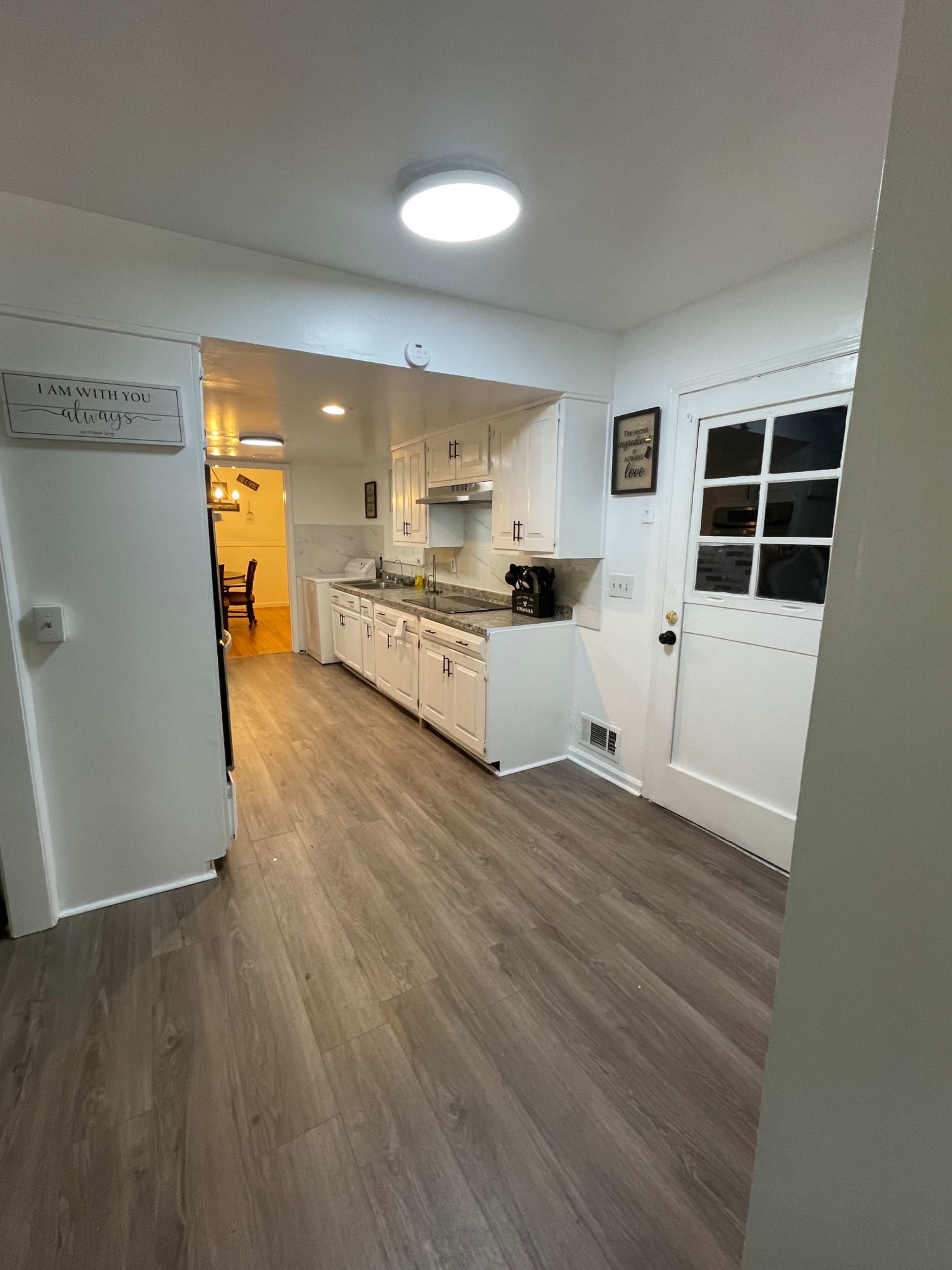 The image shows a spacious kitchen with white cabinets, a sink, and a doorway leading to a well-lit dining area.