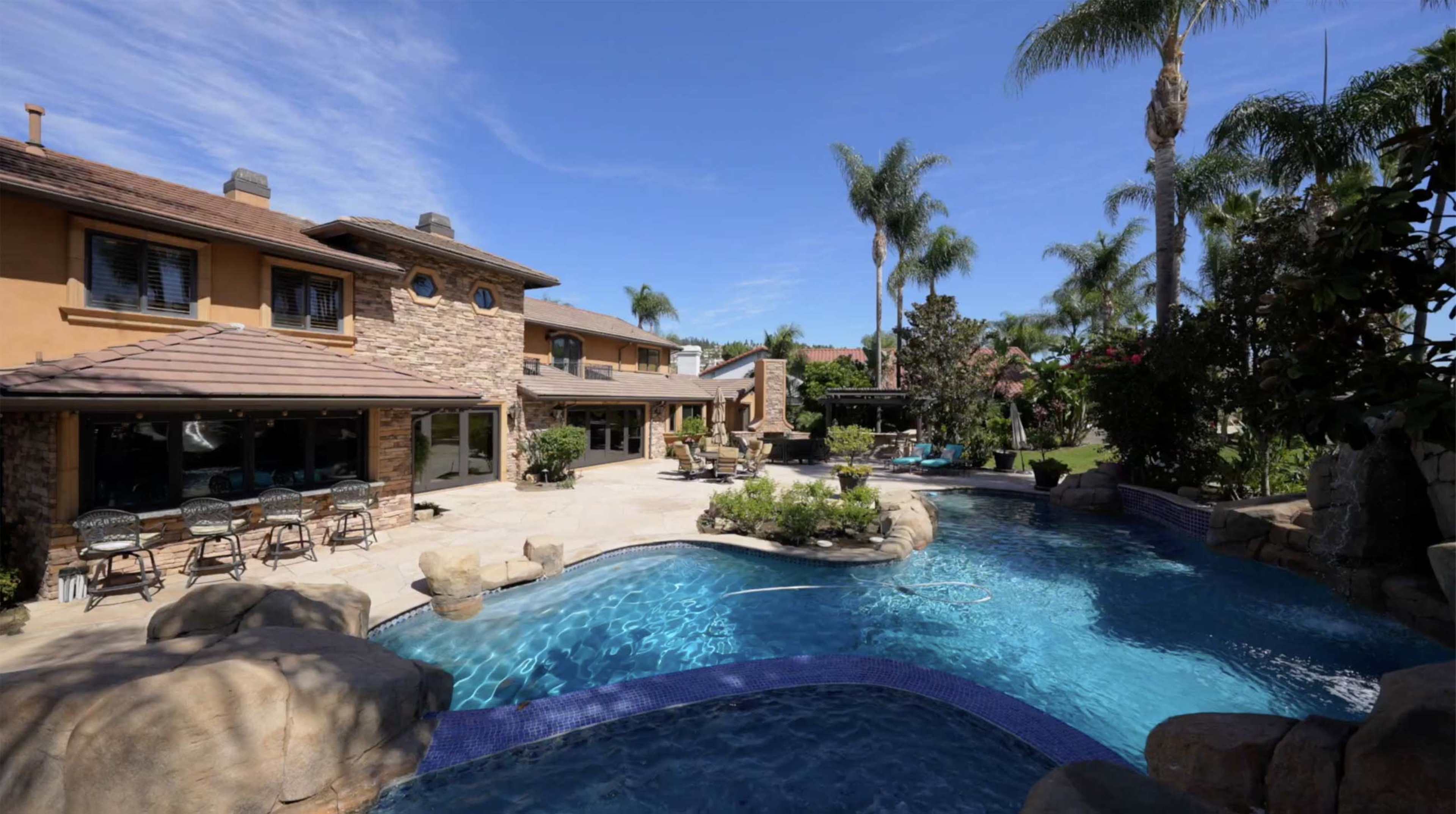 A spacious backyard with a large swimming pool, stone accents, and palm trees, adjacent to a multi-level house.