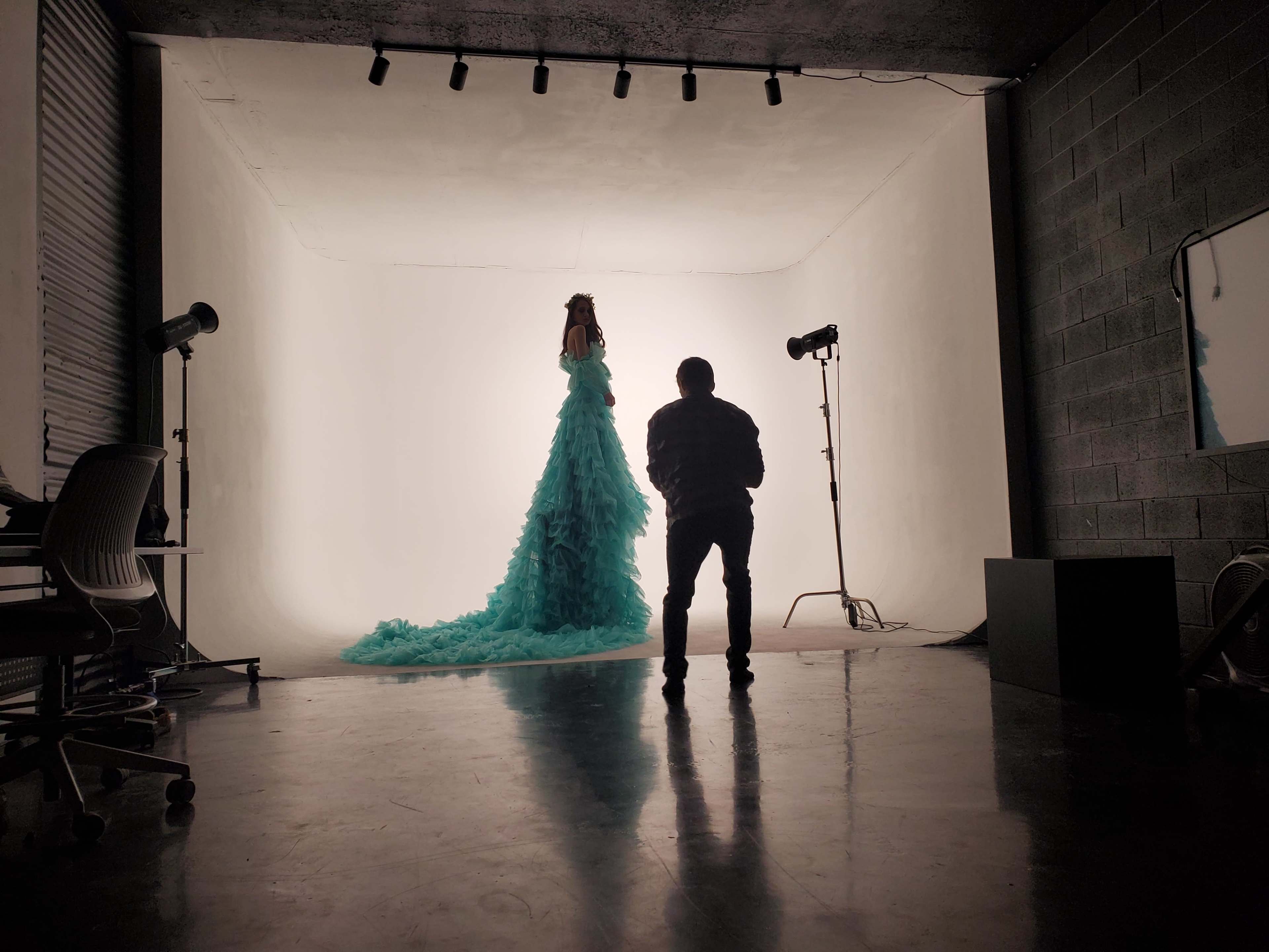 A model in a long, textured turquoise gown stands against a brightly lit backdrop while a photographer captures the scene in a studio.