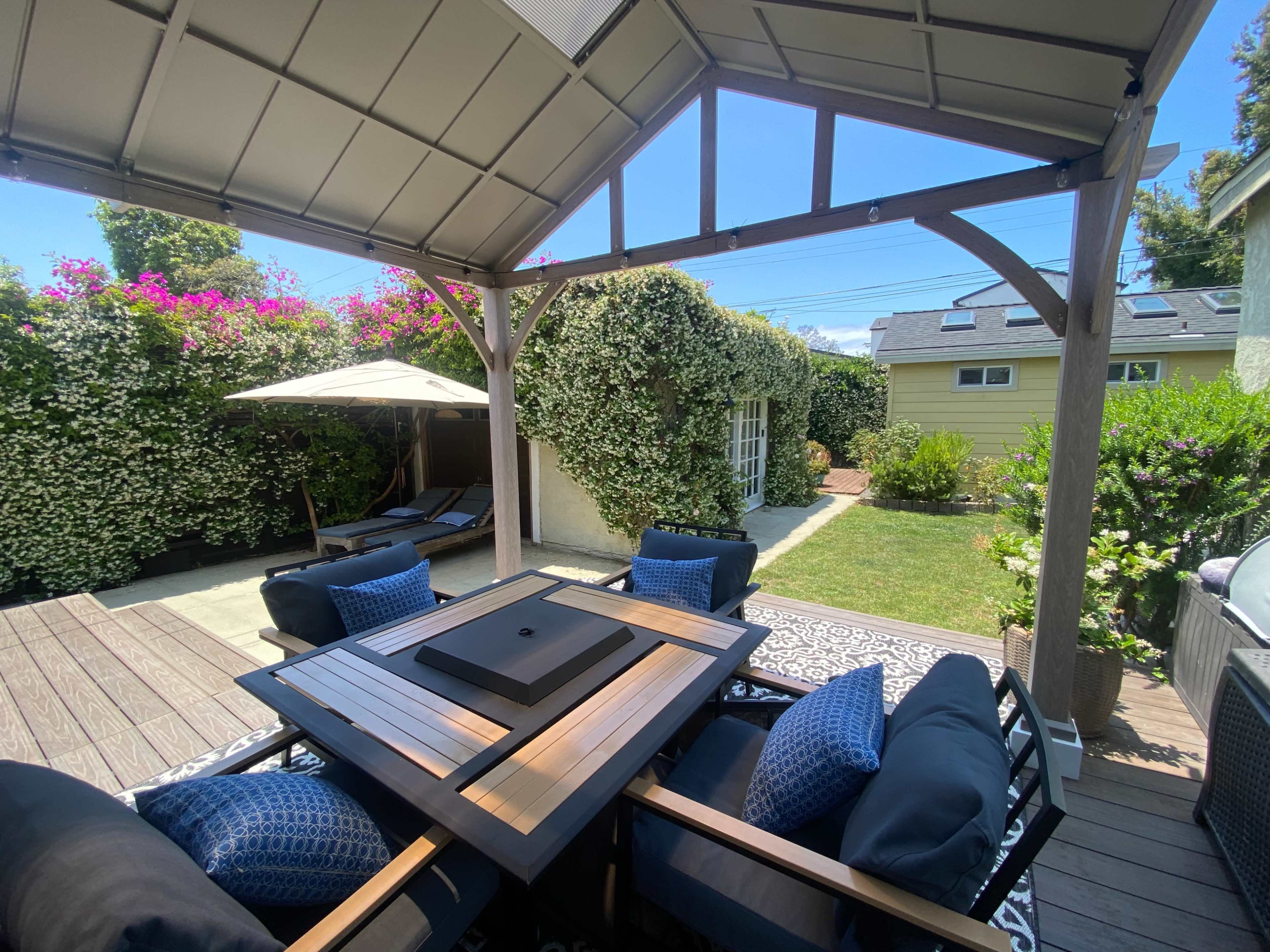 A shaded outdoor patio features a wooden table with a fire pit surrounded by four chairs, overlooking a landscaped yard with nearby lounge chairs and a sun umbrella.