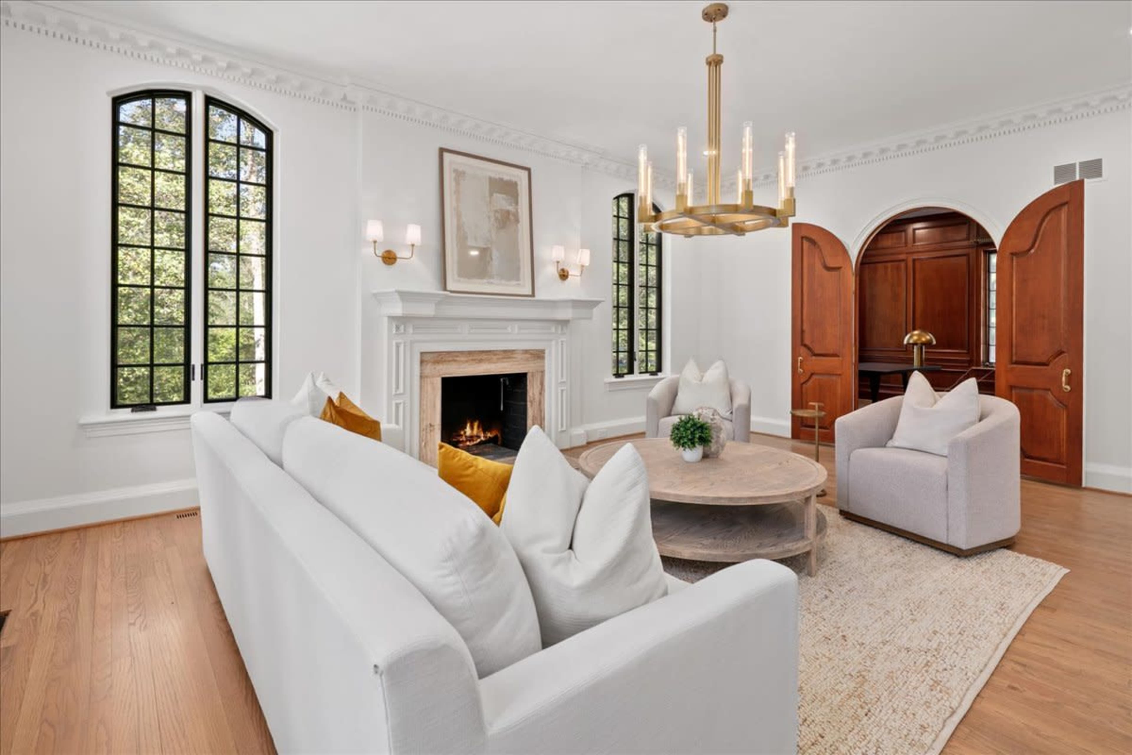 The living room features a white sofa, two armchairs, a circular coffee table, a fireplace with a stone surround, and large windows allowing natural light to enter.