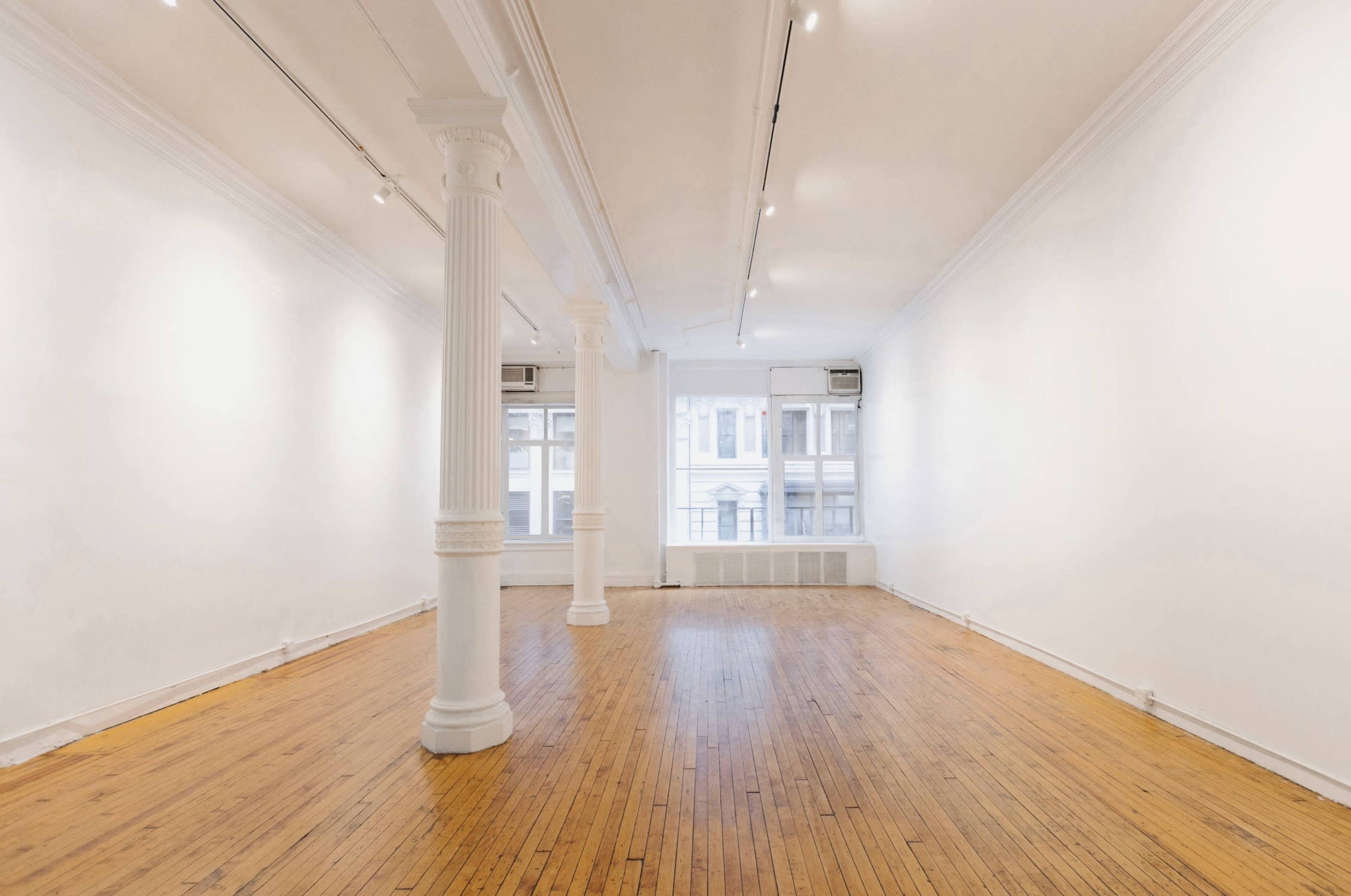 The image shows a spacious, empty room with hardwood floors, white walls, and several large windows that allow natural light to enter.
