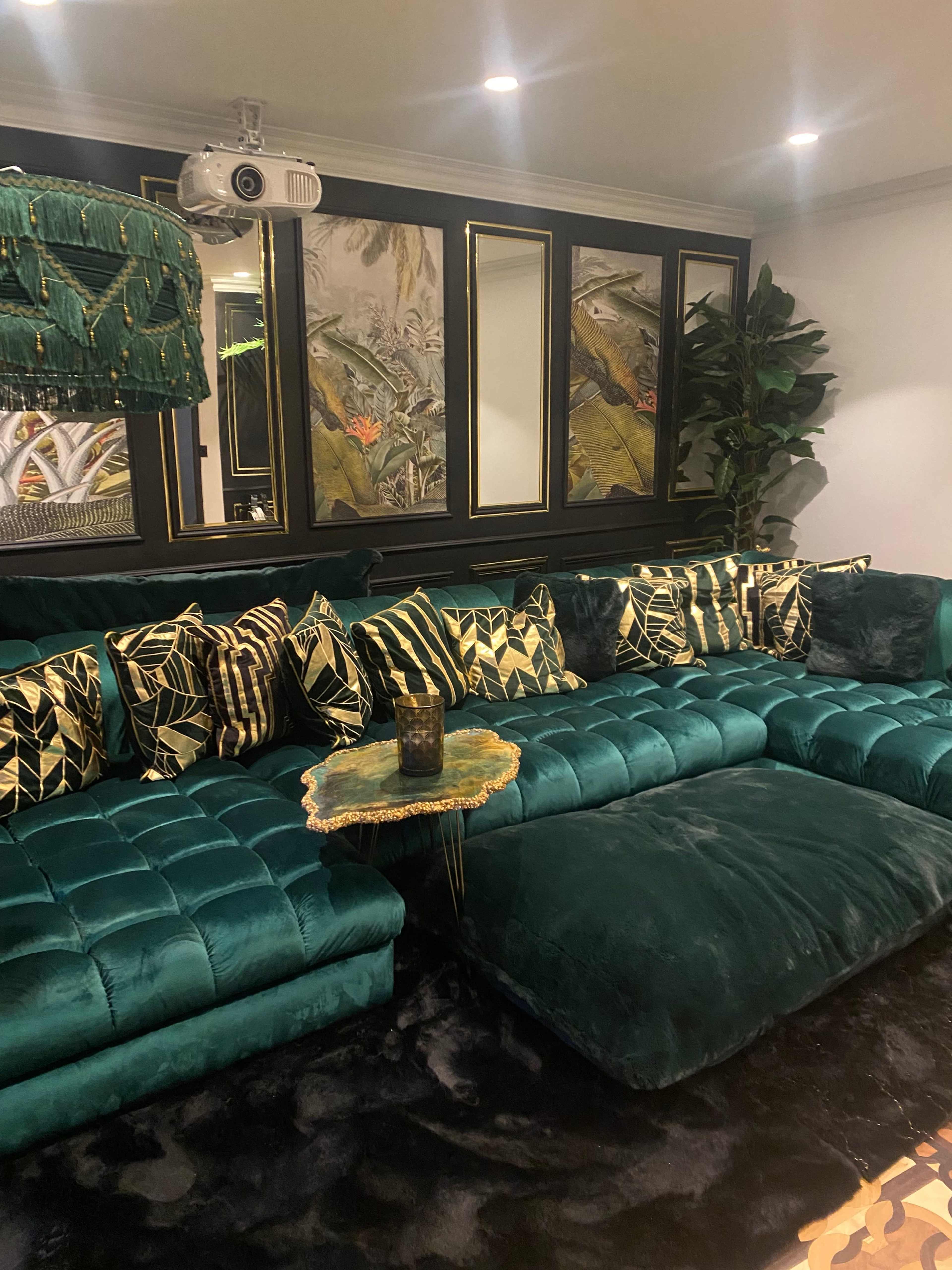 A luxurious living room featuring a green velvet sectional sofa adorned with patterned cushions, a small decorative table, and a projector mounted on the ceiling.