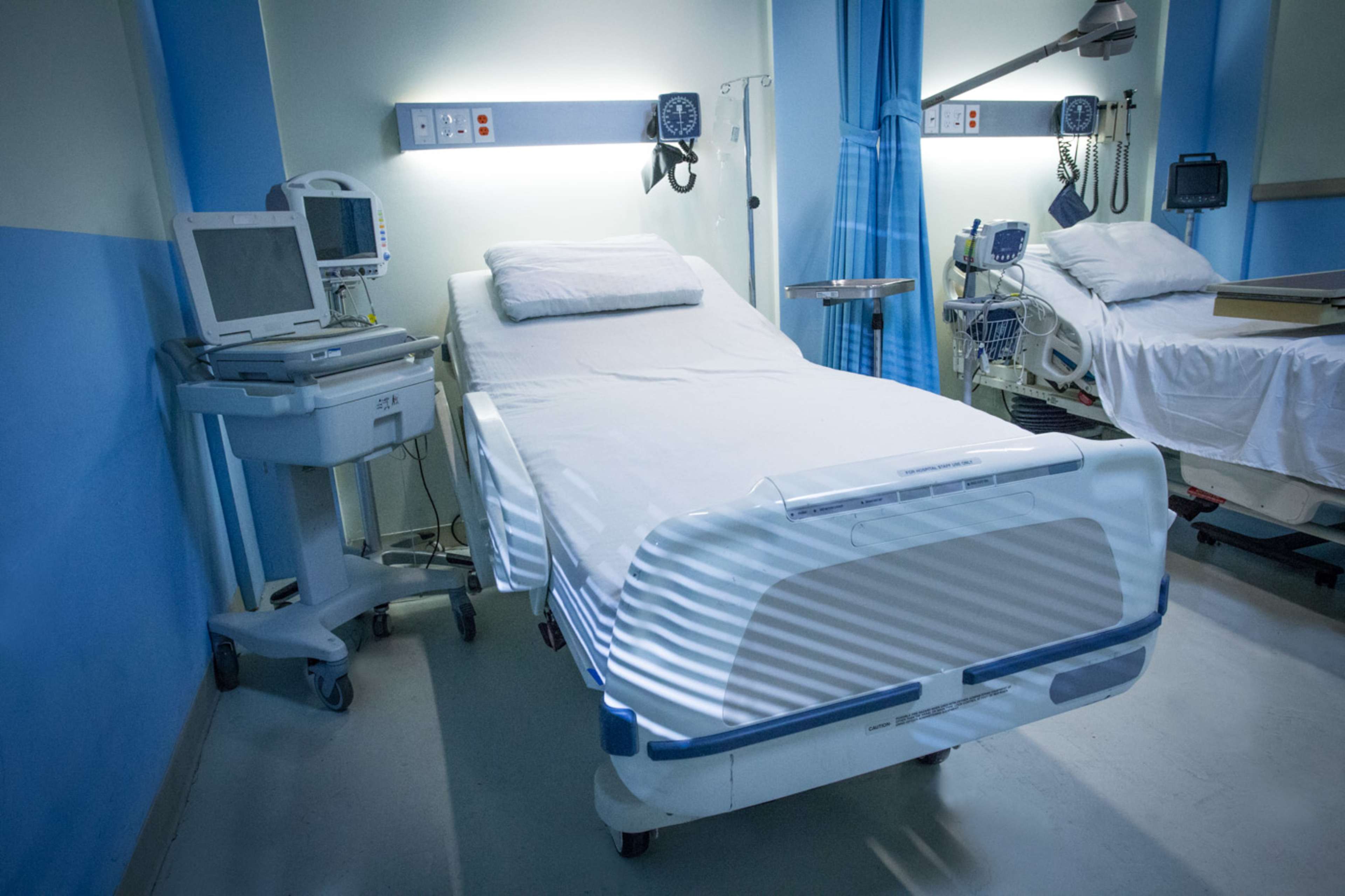A hospital room with two empty beds, medical equipment beside them, and blue and green walls.
