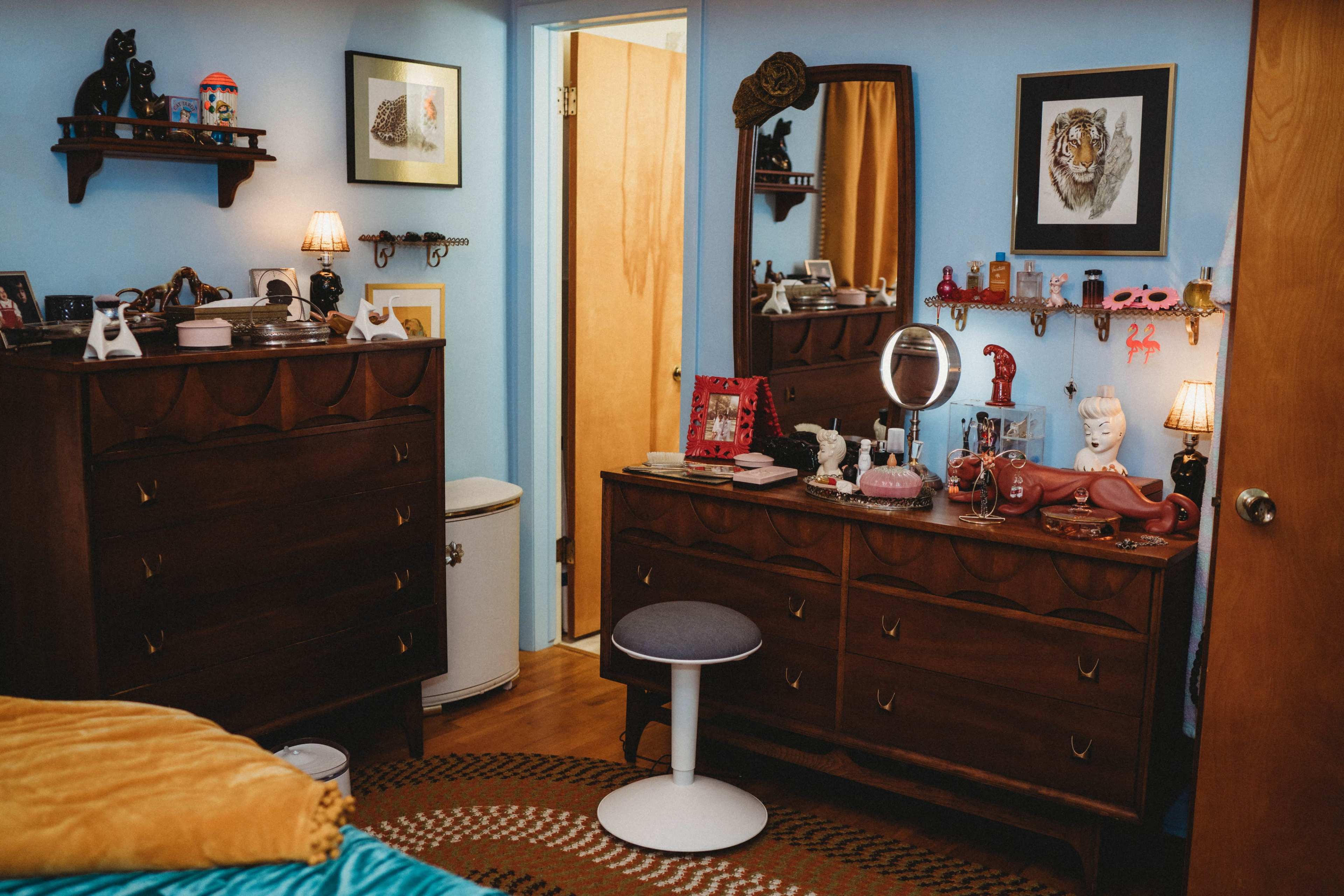 The image shows a cozy bedroom featuring a wooden dresser and mirror, a stool, and various decorative items against a blue wall.