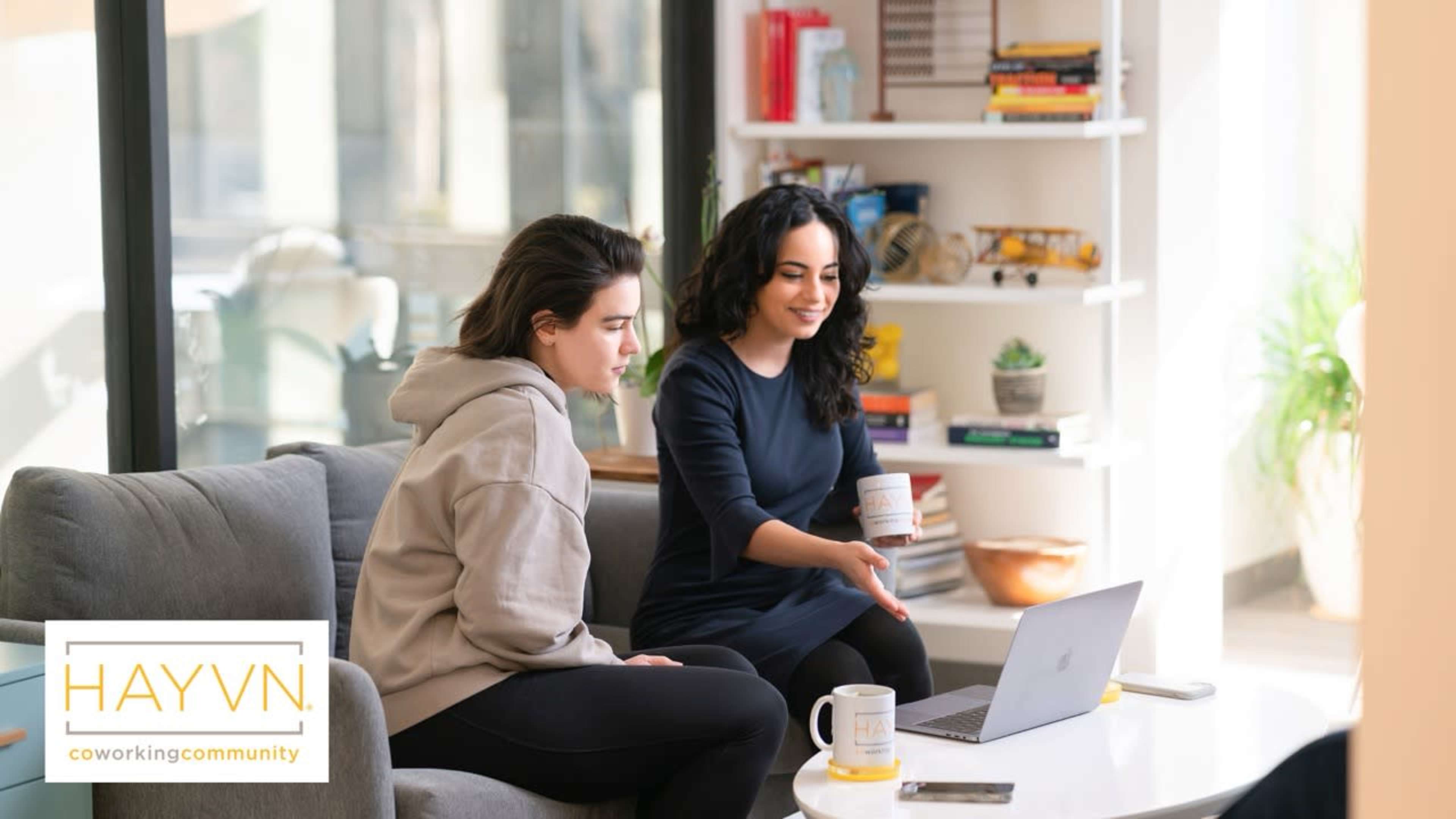 Two women are sitting on a couch next to a laptop, discussing something while one gestures towards the screen in a bright, modern coworking space.