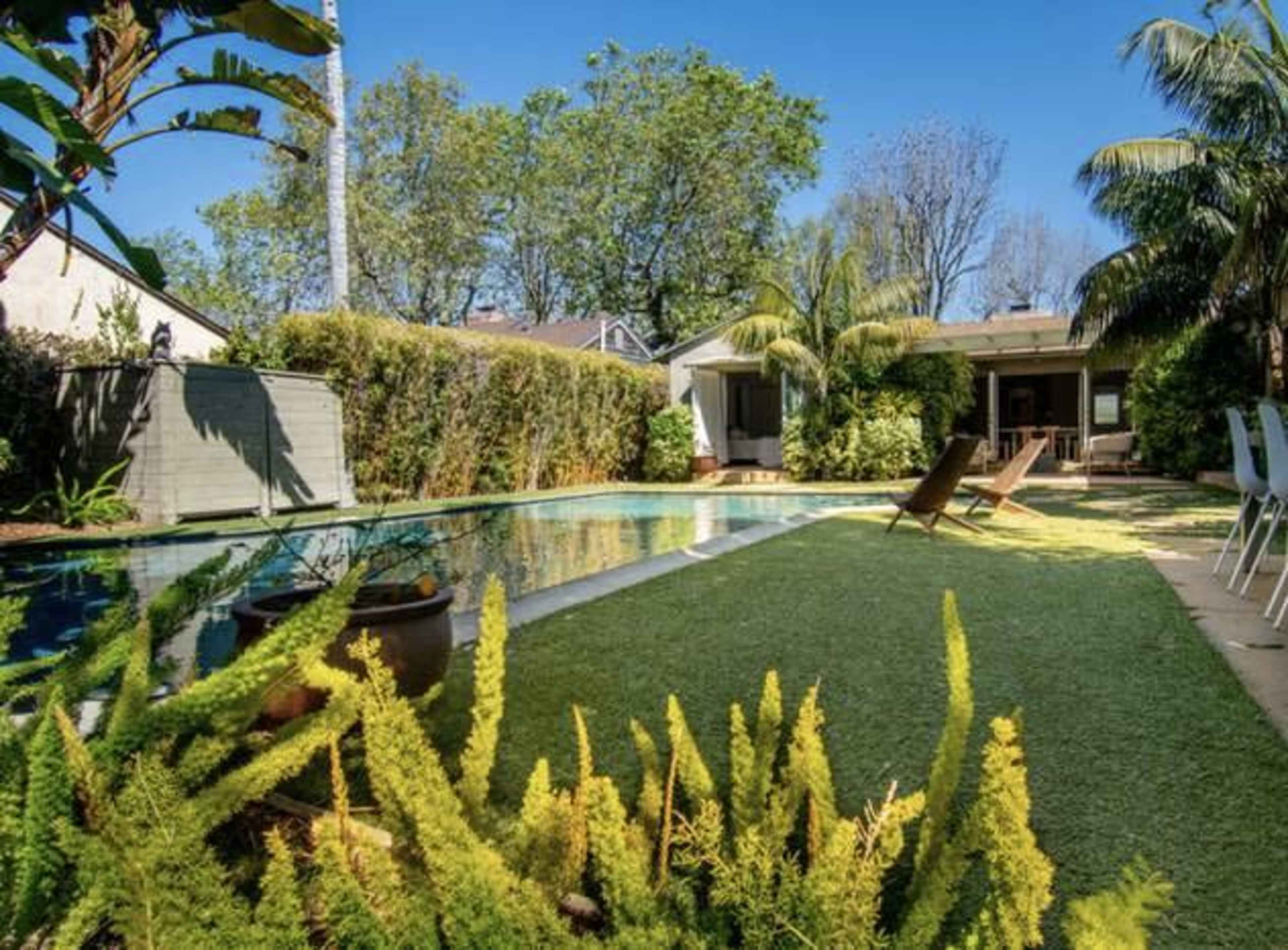 A backyard with a swimming pool surrounded by greenery and lounge chairs, featuring a house in the background.