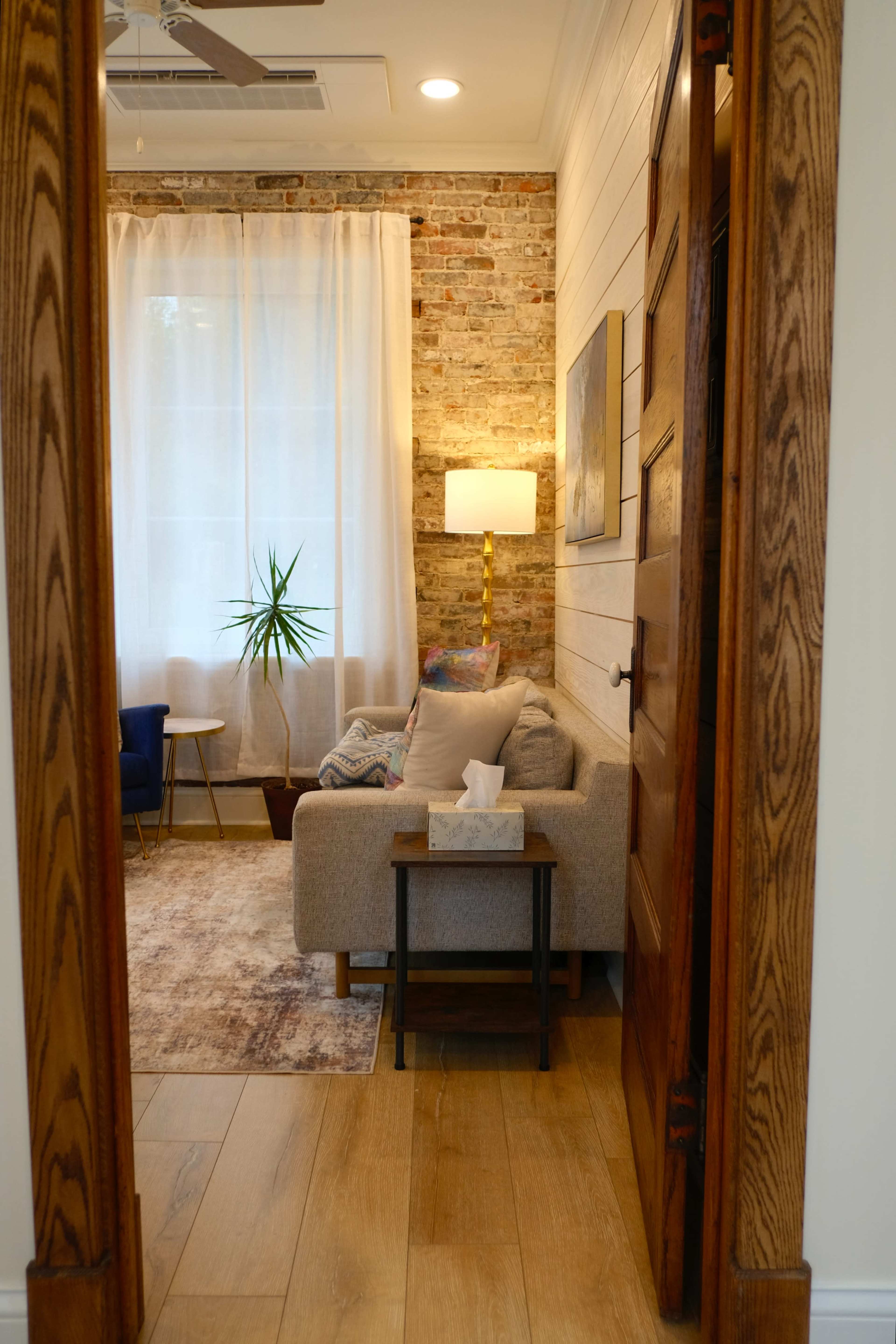 A cozy living room is visible through an open door, featuring a sofa, a potted plant, a lamp, and a large window with sheer curtains.