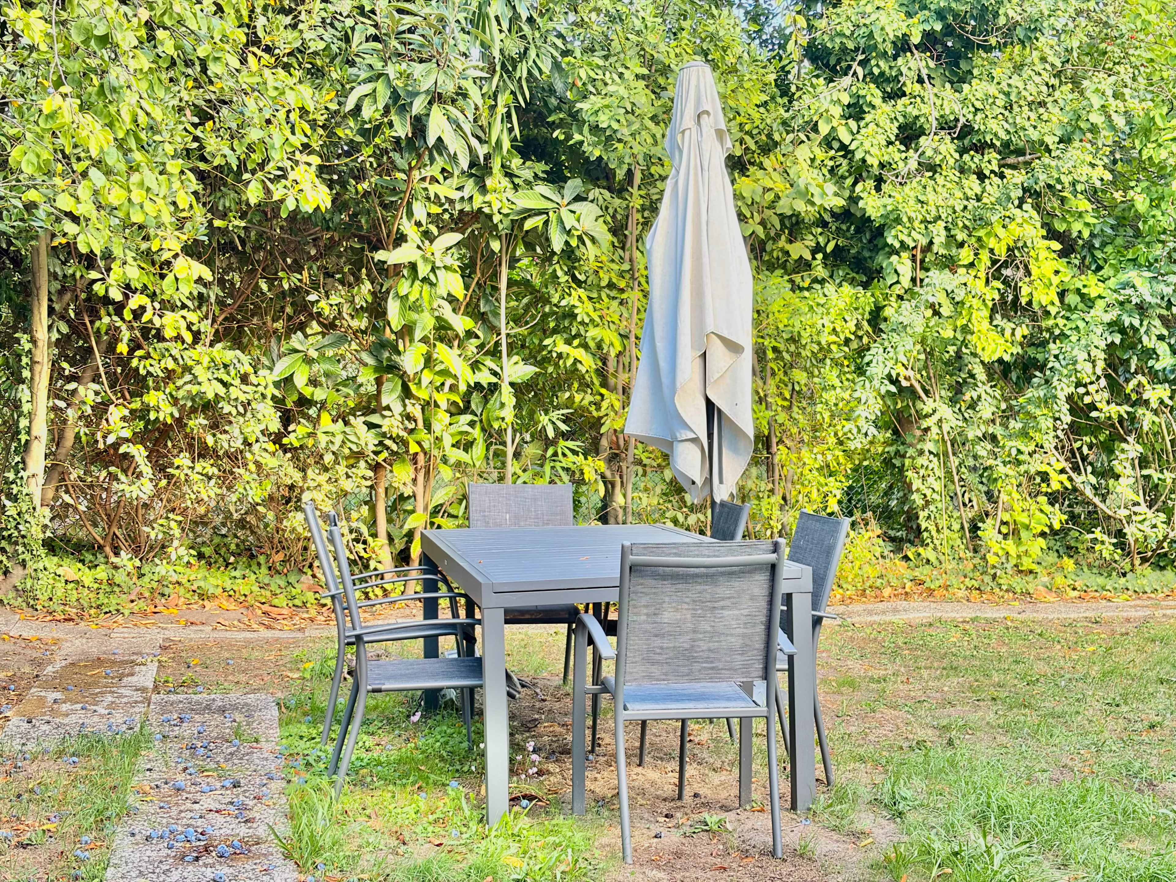 A gray patio table with four chairs and an umbrella stands in a grassy area surrounded by greenery.