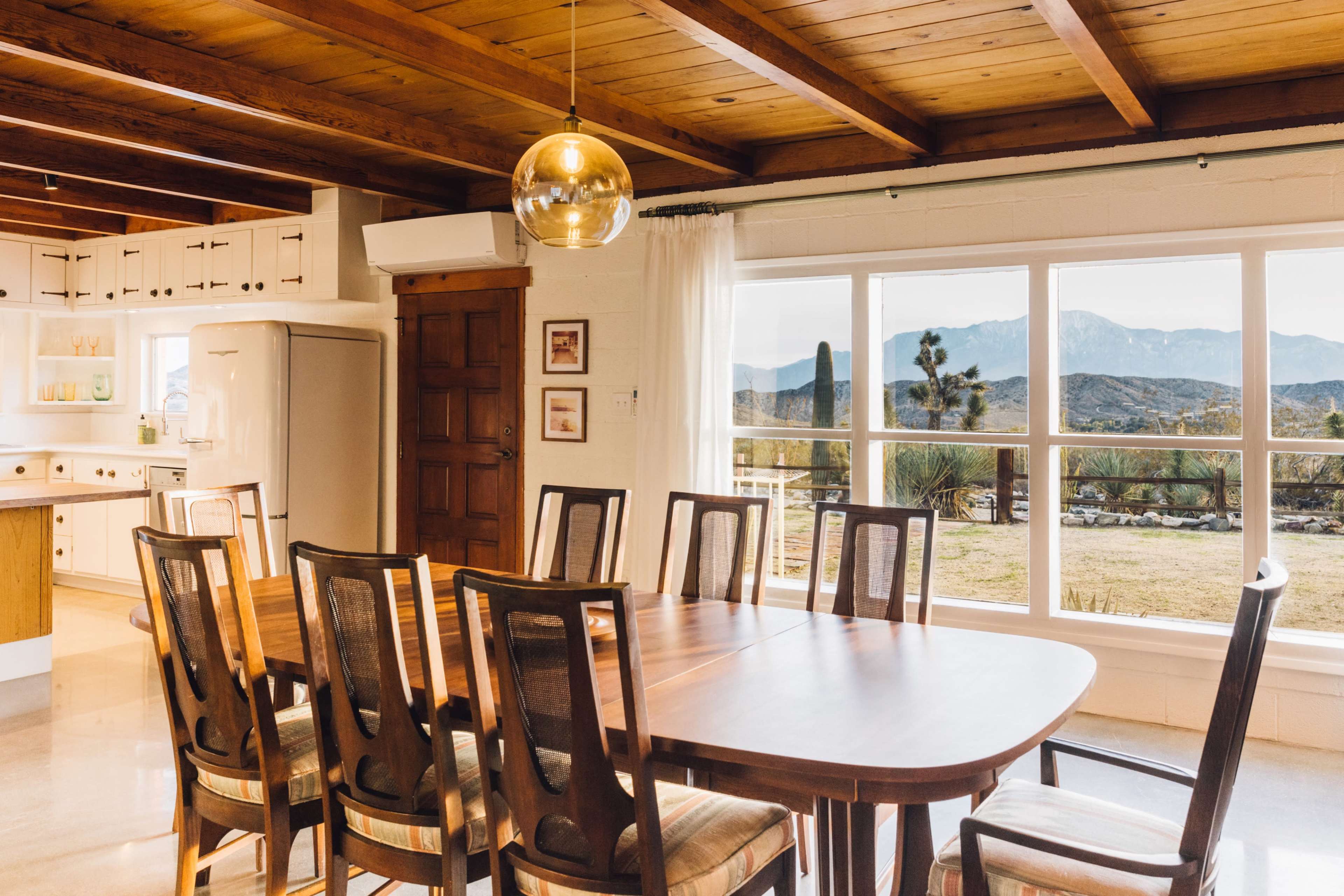 A wooden dining table with chairs is set in a brightly lit room featuring large windows that overlook a mountainous landscape.