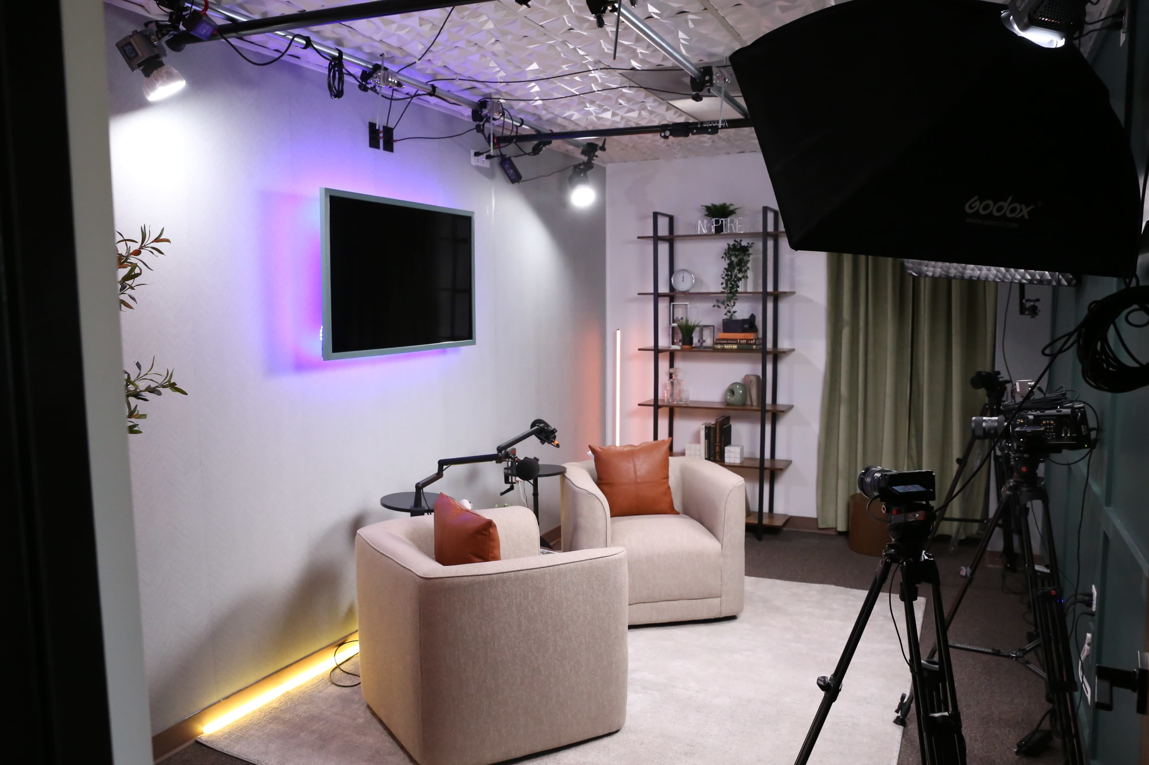The image shows a well-equipped studio space with two beige chairs, a television mounted on the wall, and various lighting and camera equipment set up for filming.
