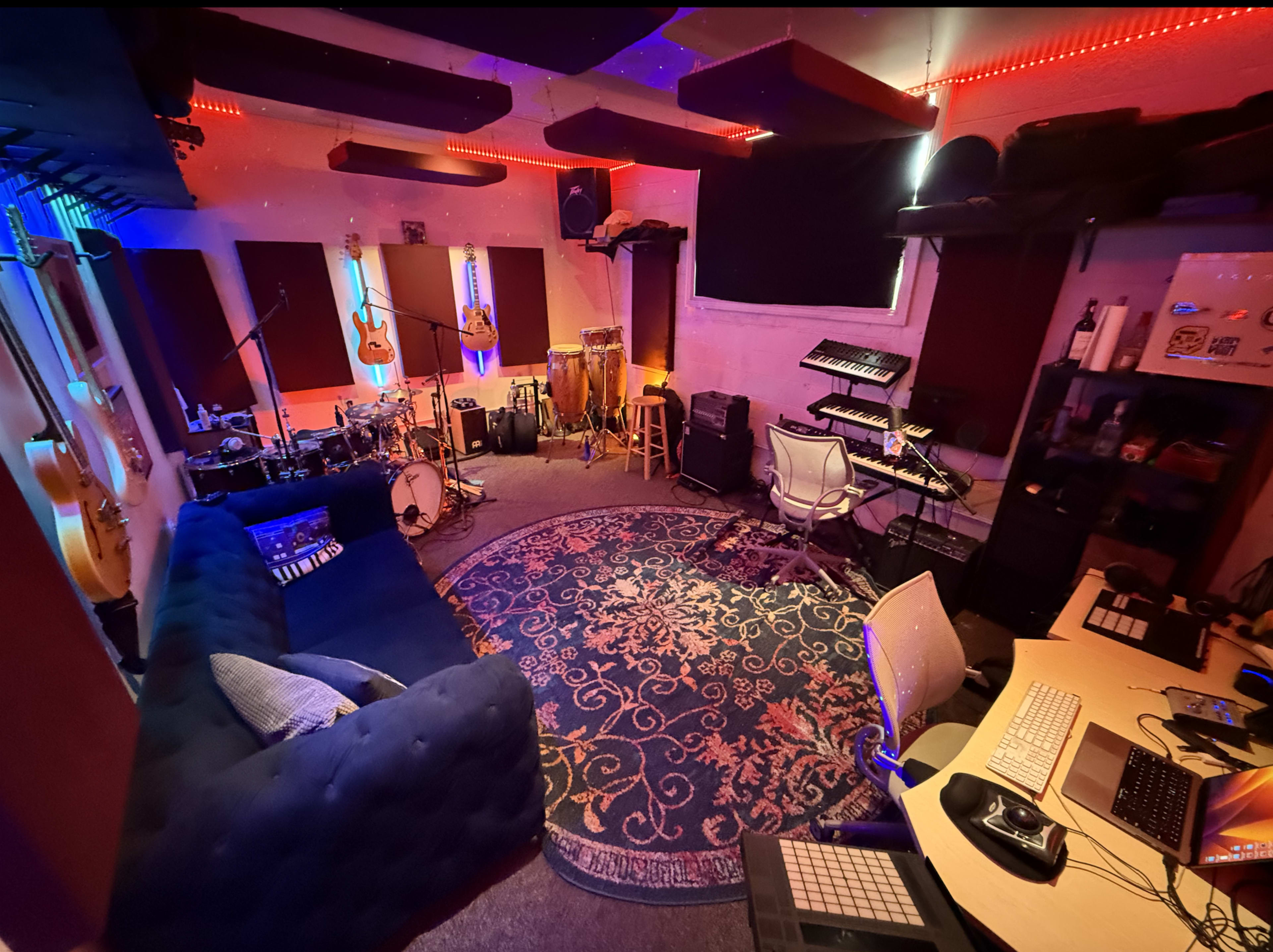 The image shows a music recording studio with various instruments, including guitars and percussion, a cozy couch, and electronic equipment arranged around a central area with a patterned rug.