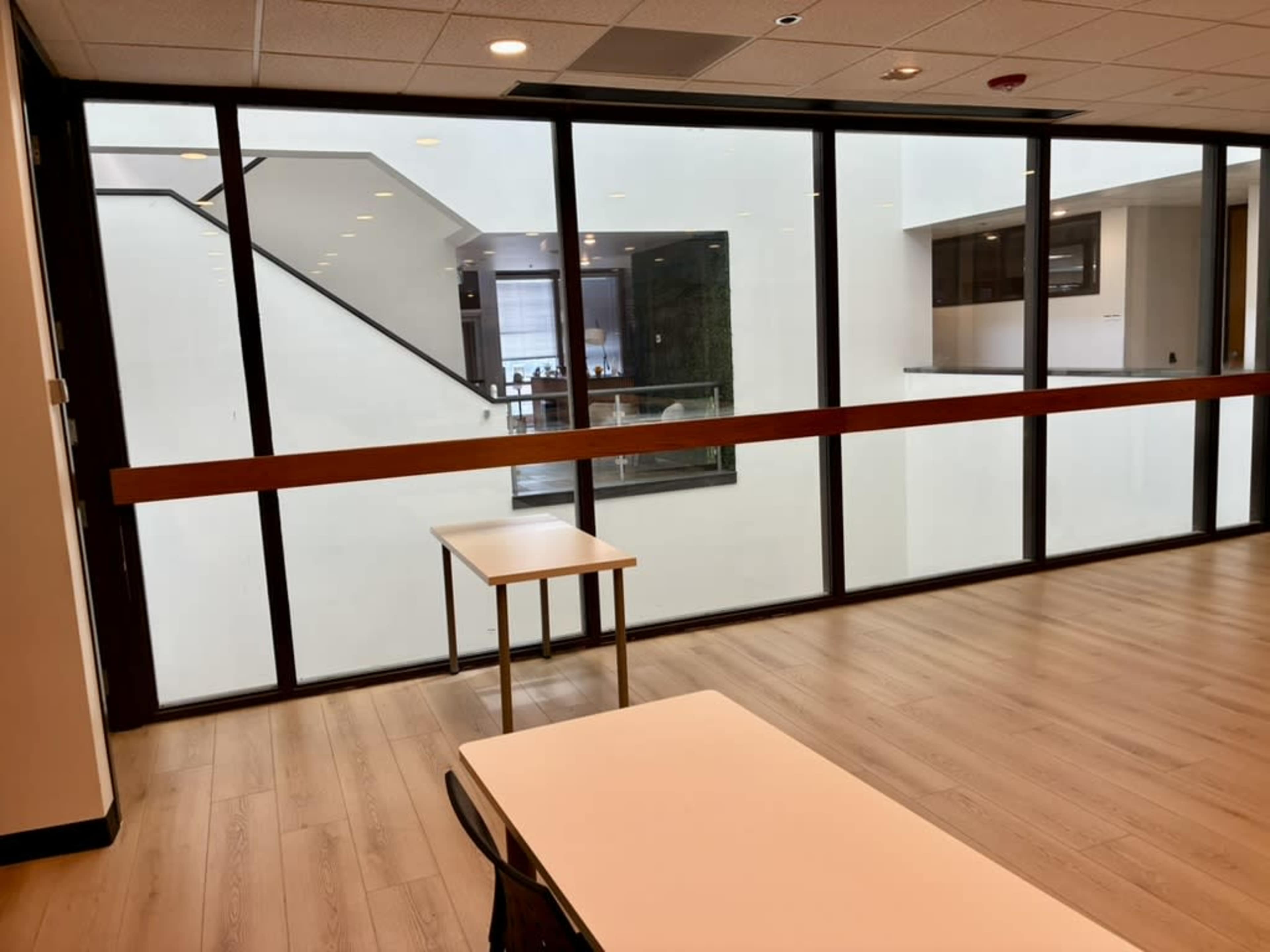 The image shows an interior space with wooden flooring, two empty tables, and large glass windows overlooking a modern, open atrium area.