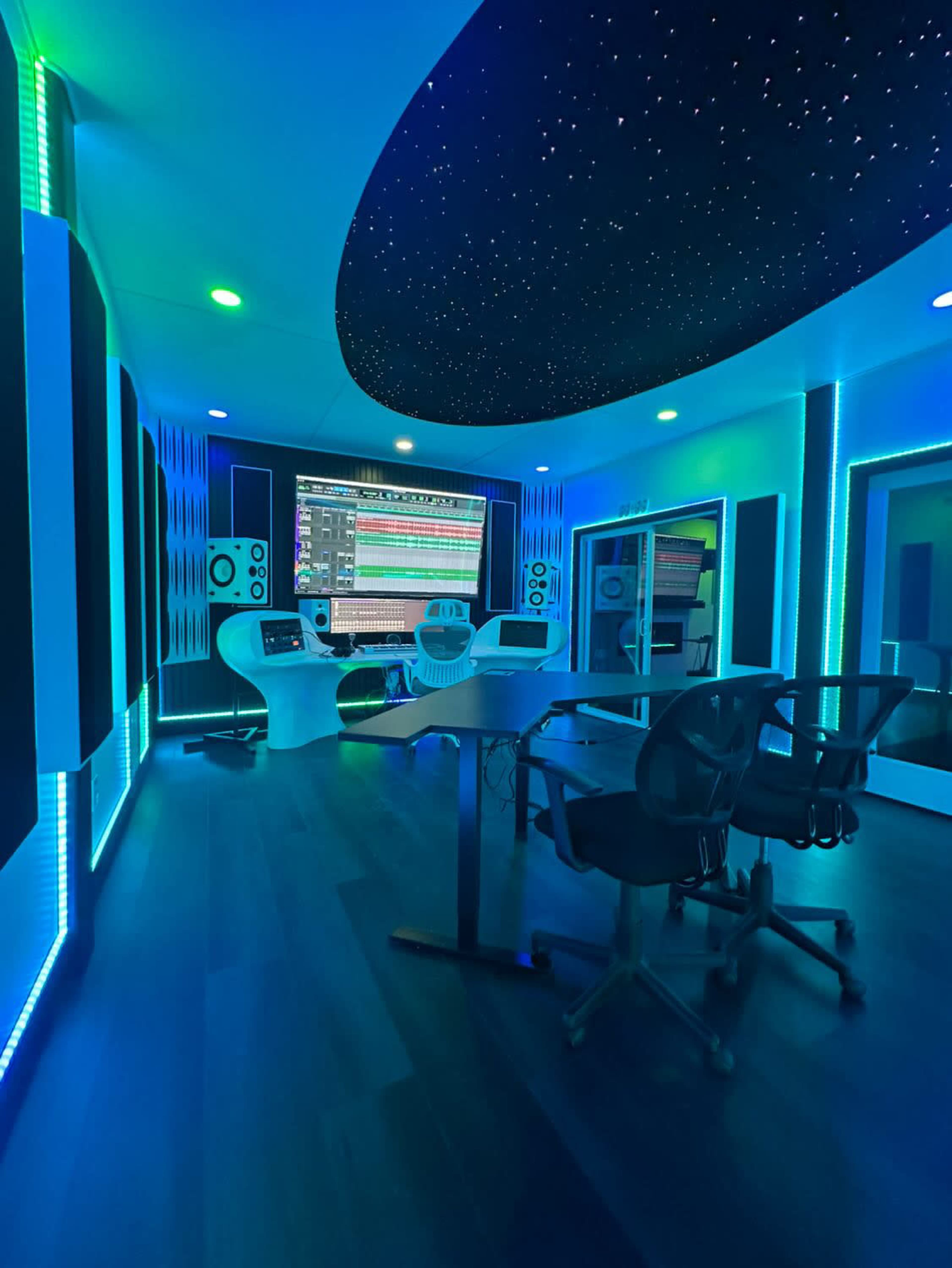 The image shows a modern music studio with blue and green LED lighting, a large central mixing console, and soundproofing panels on the walls.