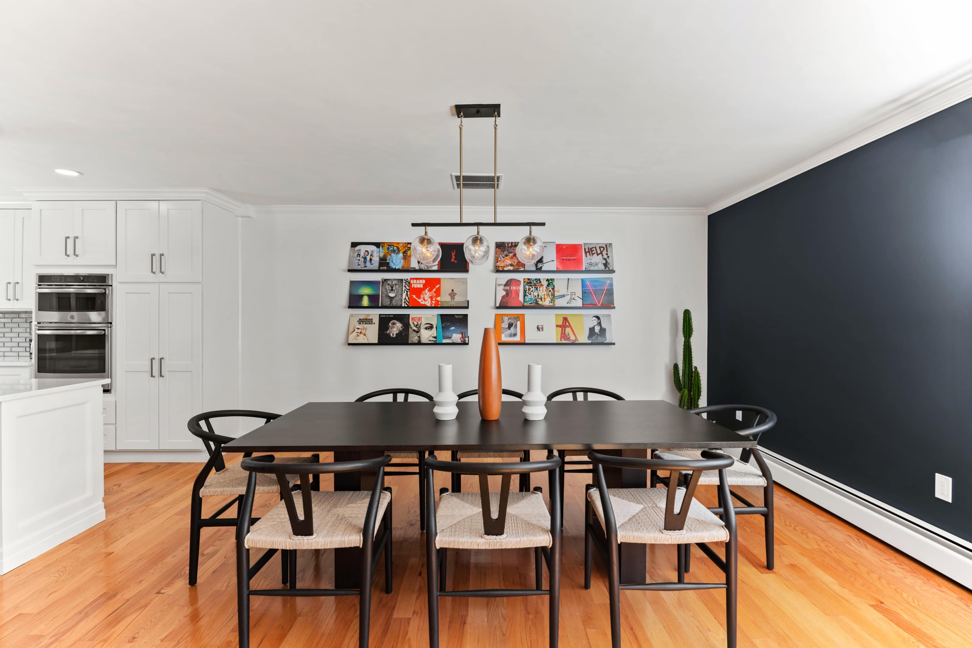 A dining room features a long black table surrounded by eight wooden chairs, with a wall-mounted shelf displaying colorful magazines.