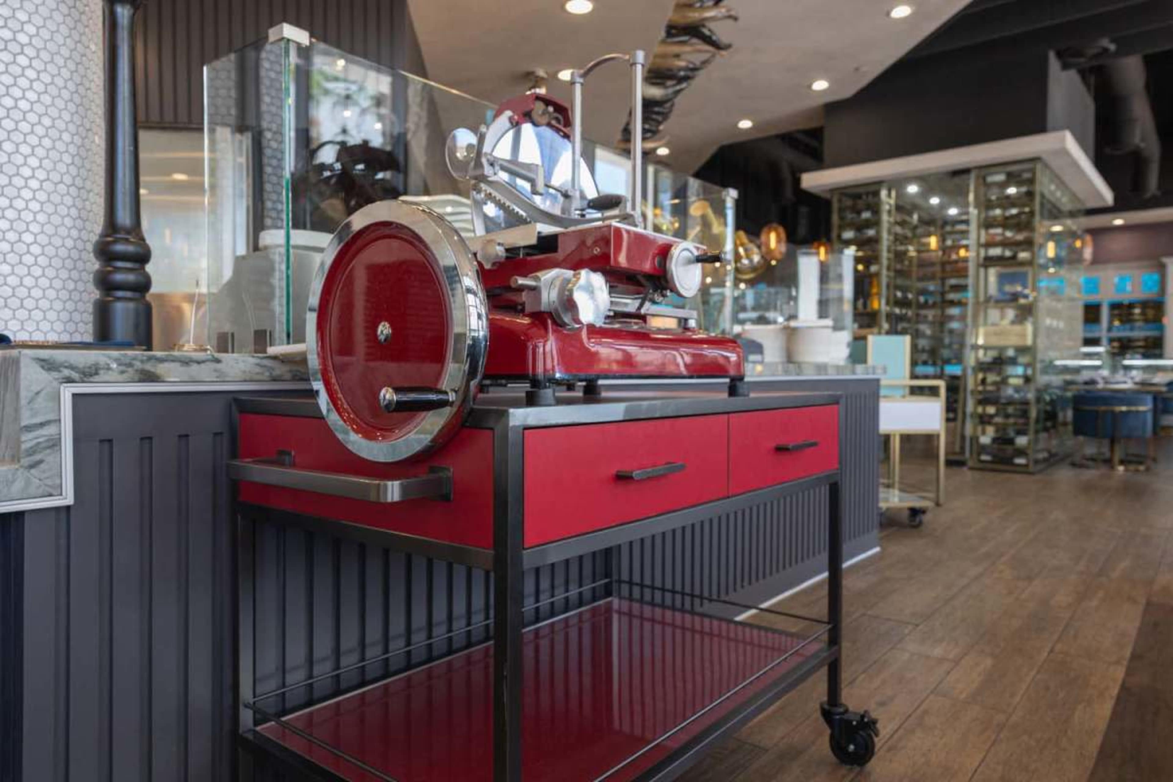 A red meat slicer is displayed on a mobile black cart within a modern commercial kitchen setting.