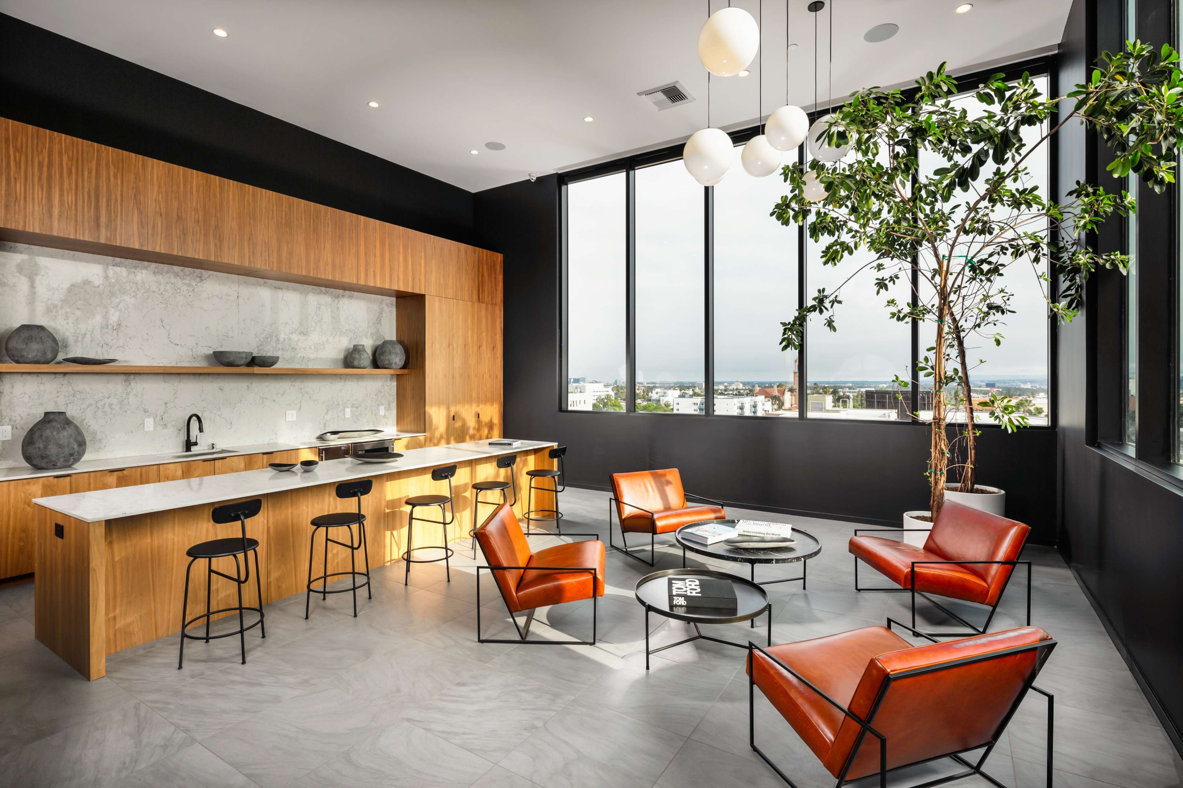 A modern lounge area features a wooden cabinetry wall, a marble countertop, four orange chairs, a round glass coffee table, and large windows displaying a cloudy view.