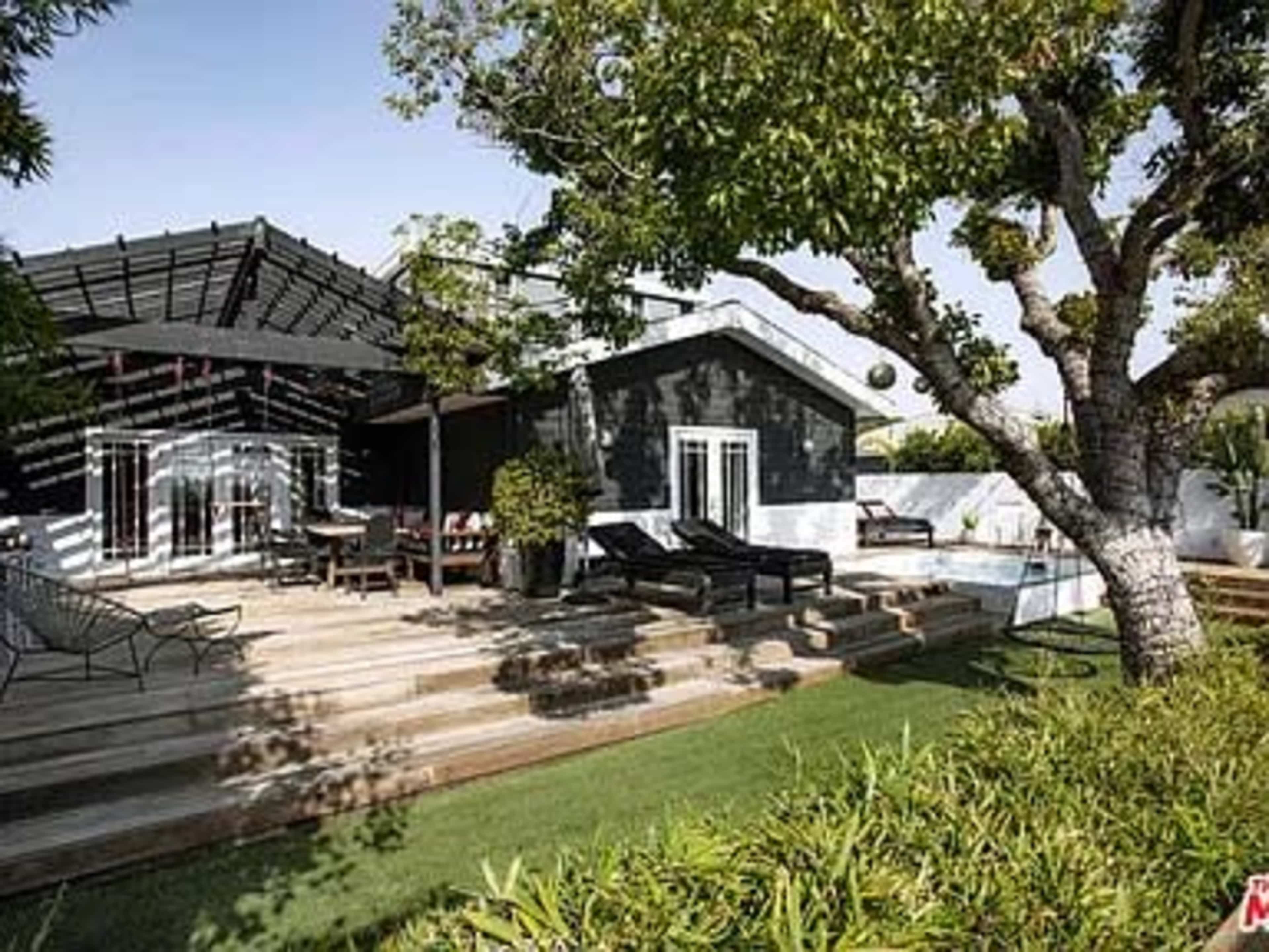 A modern house with a black exterior, surrounded by a wooden deck, lounge furniture, and landscaped greenery.