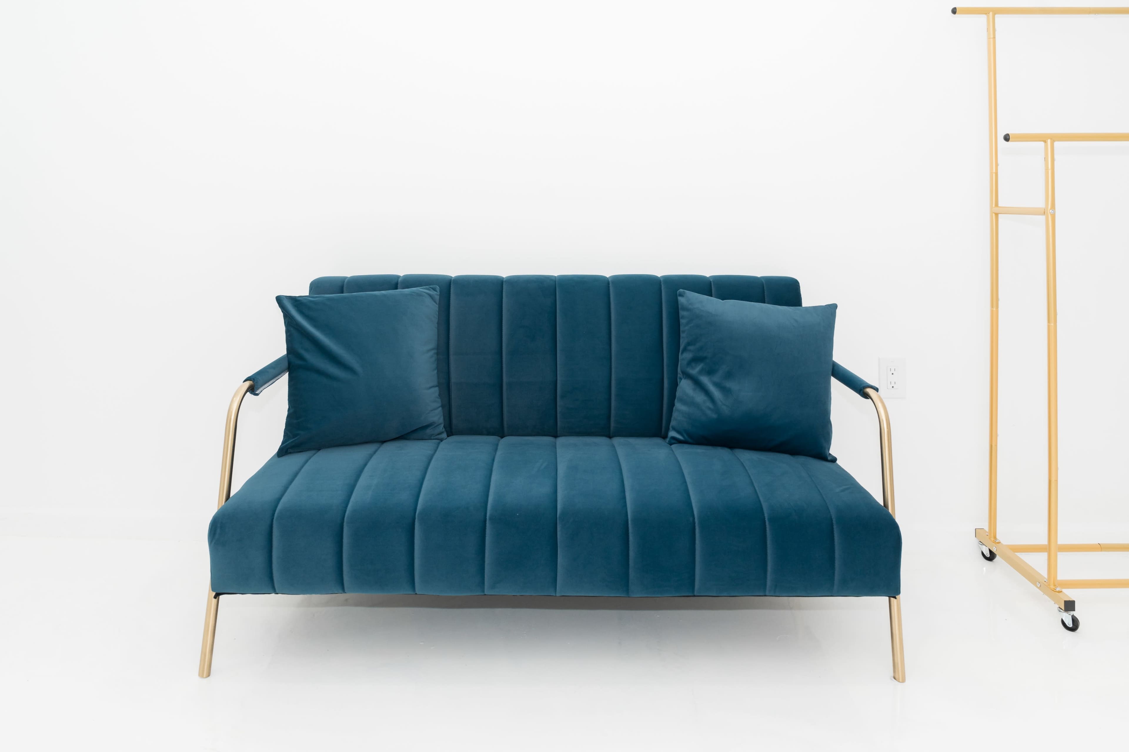 A teal velvet sofa with gold metal arms and two matching cushions is positioned against a white wall.