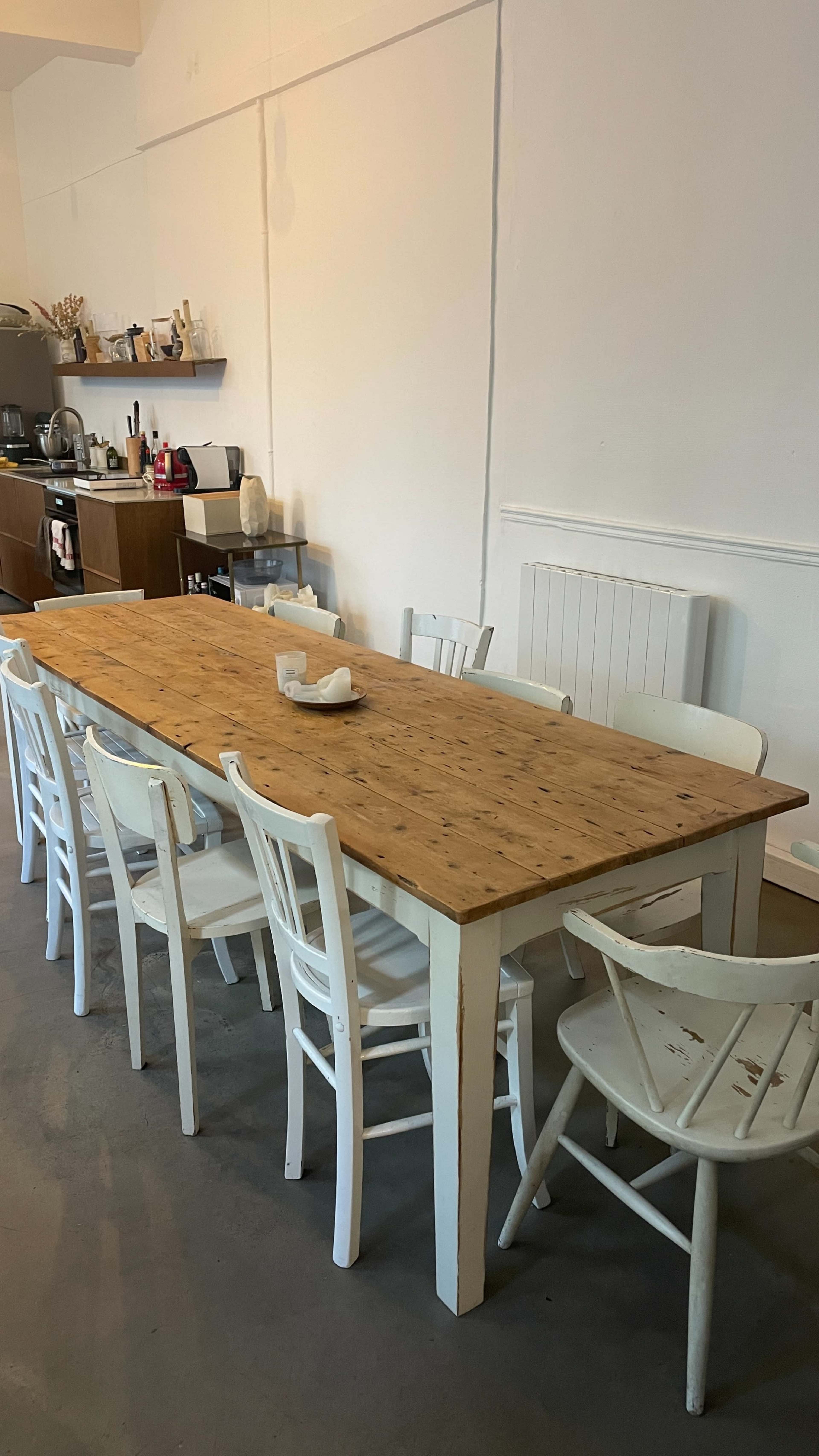 A long wooden table with a light-colored finish is surrounded by several white, wooden chairs in a minimalistic interior setting.